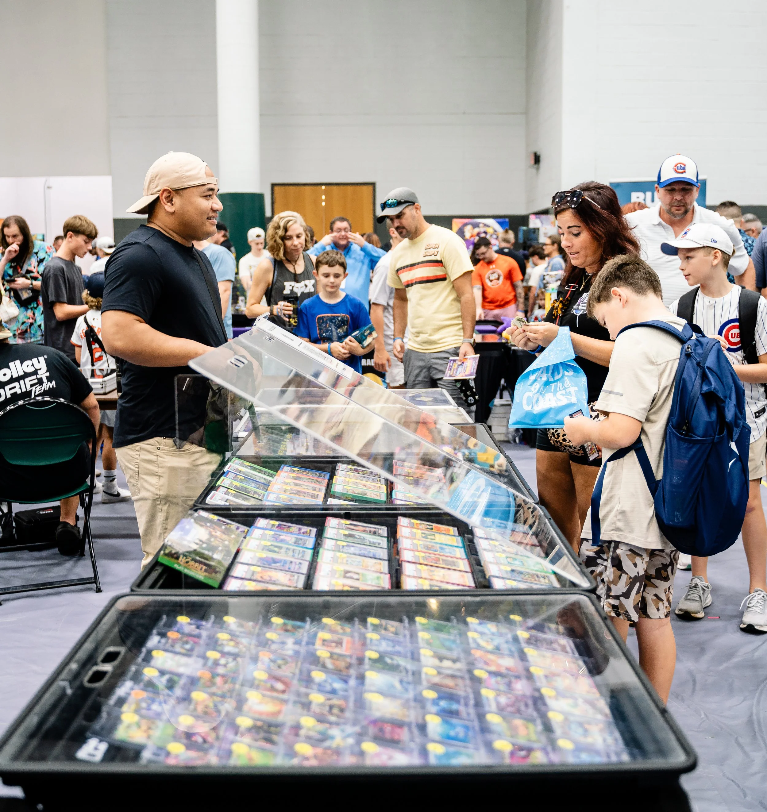 A large indoor event with people browsing tables displaying various collectible cards and toys. The foreground shows a glass display case filled with colorful toy figures and cards. People are engaged, some examining items, and others chatting. The b