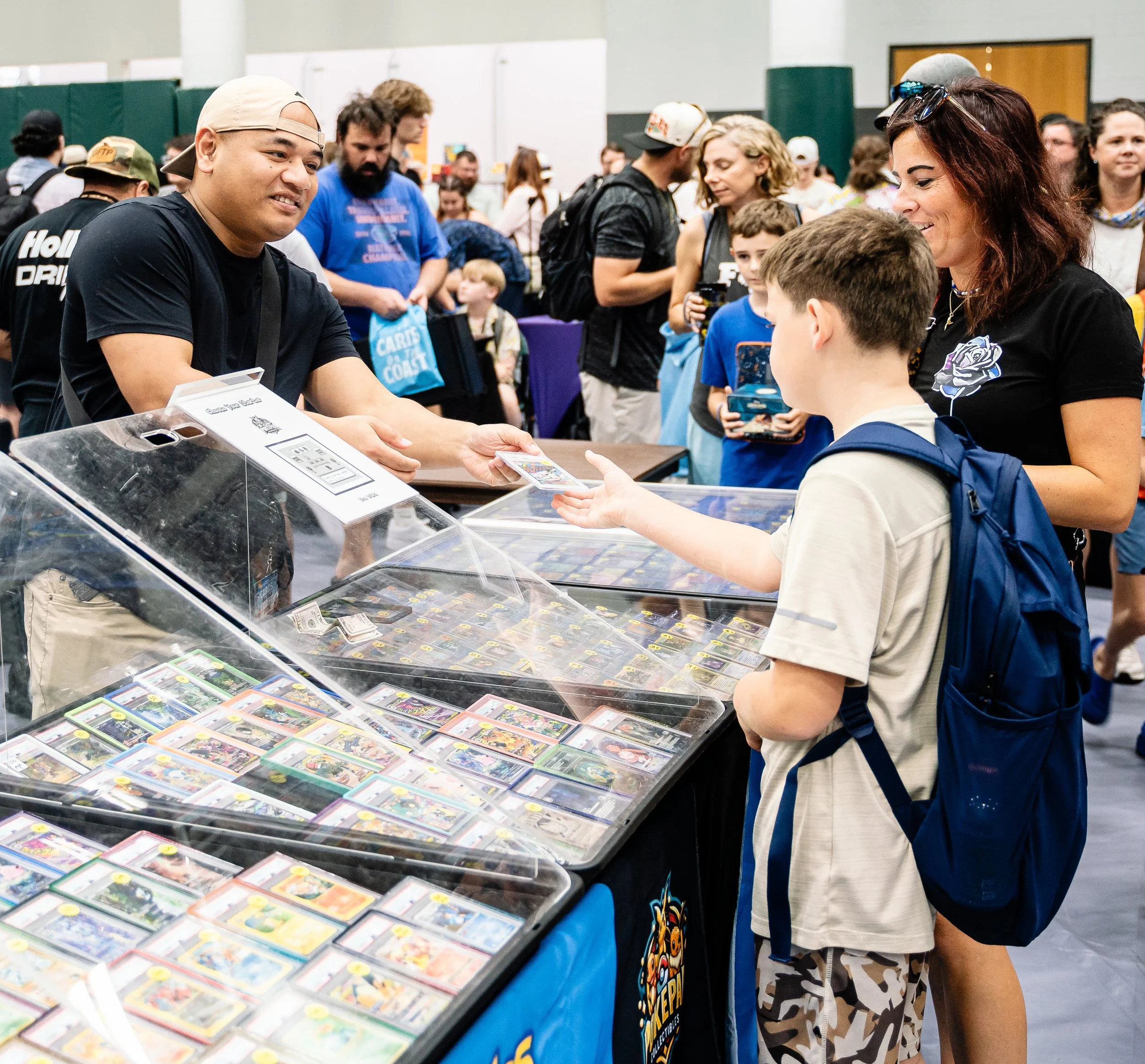 People at a convention or trade show around a display case filled with collectible cards, with some engaging in conversation and others browsing.