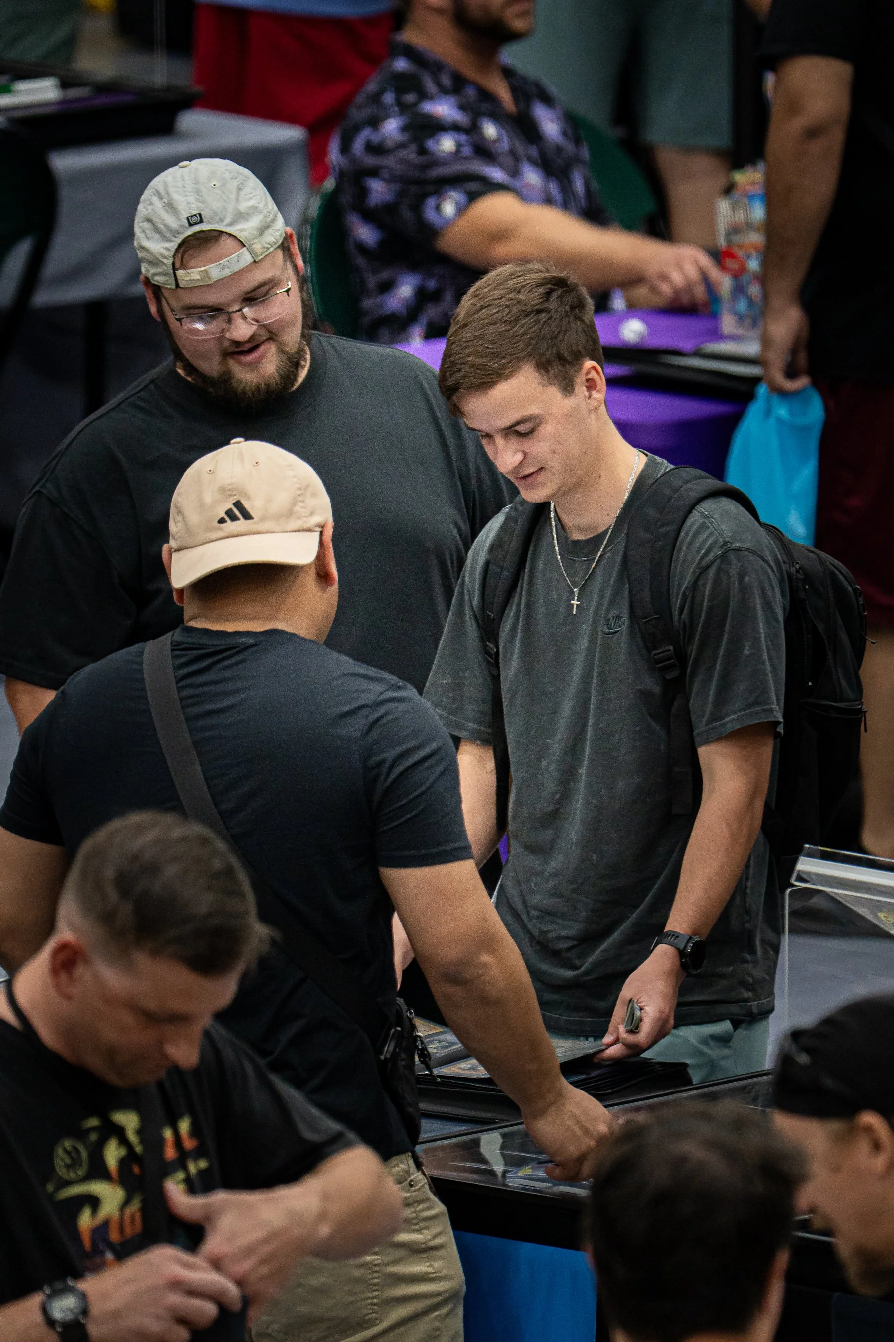 Several young men are gathered around a table at a convention or event, with various people and objects in the background. They are examining items on the table, possibly trading or selling collectibles or cards.