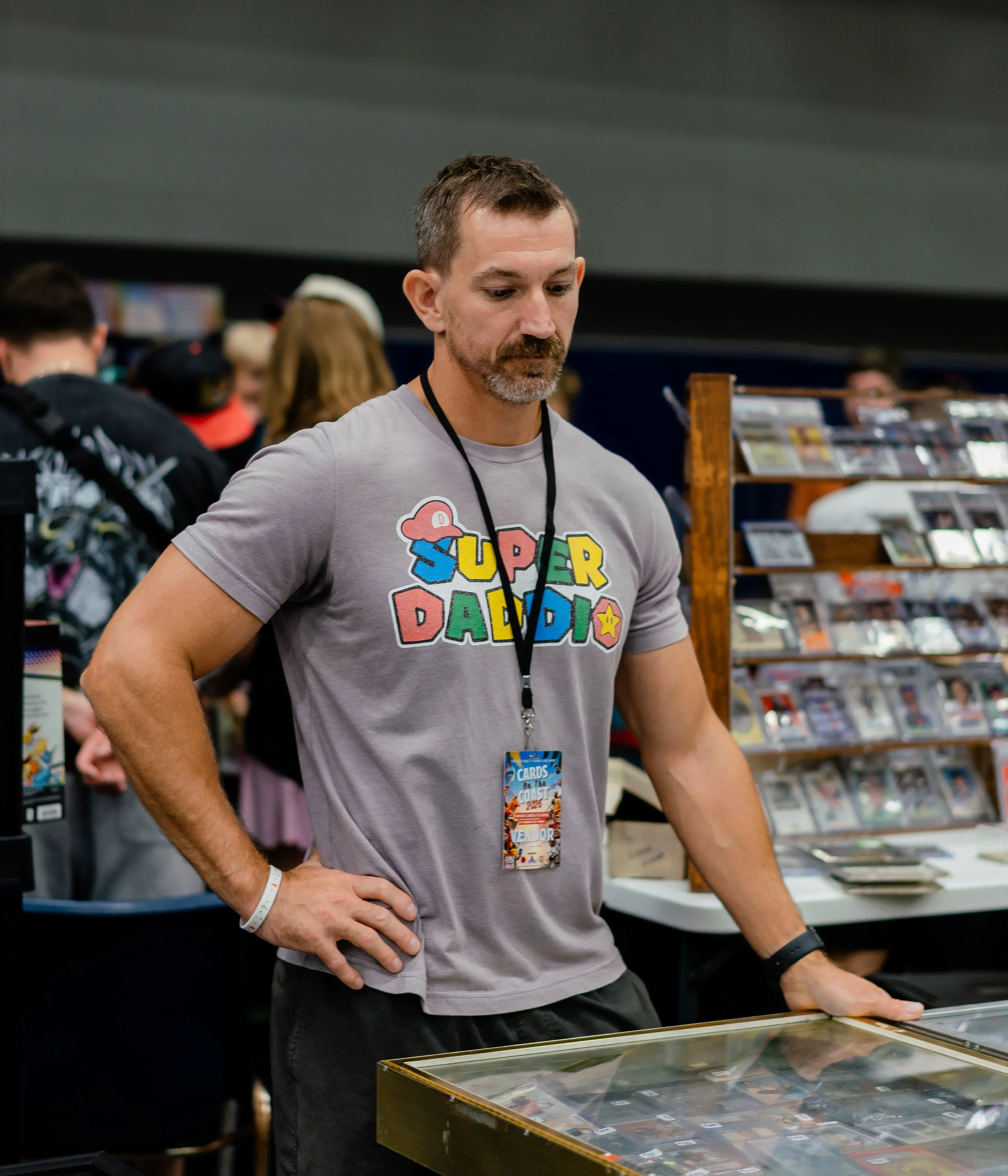 A man with short brown hair, a beard, and a grey Super Mario T-shirt browsing through a selection of trading cards at a convention or store. Others are visible in the background around display shelves.