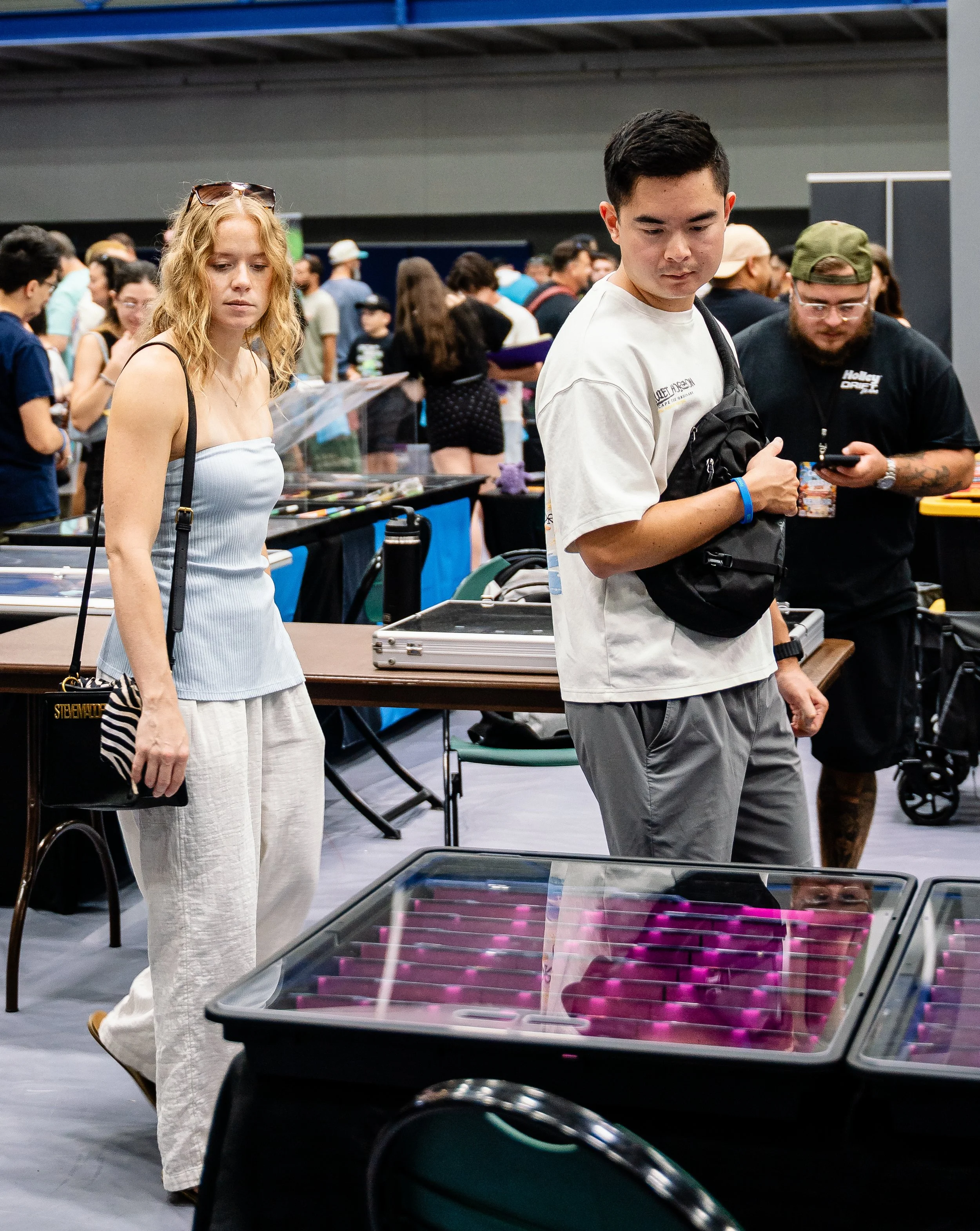 People attending an indoor event, with a table displaying pink-colored items in the foreground and a crowd of individuals in casual clothing browsing and examining items in the background.