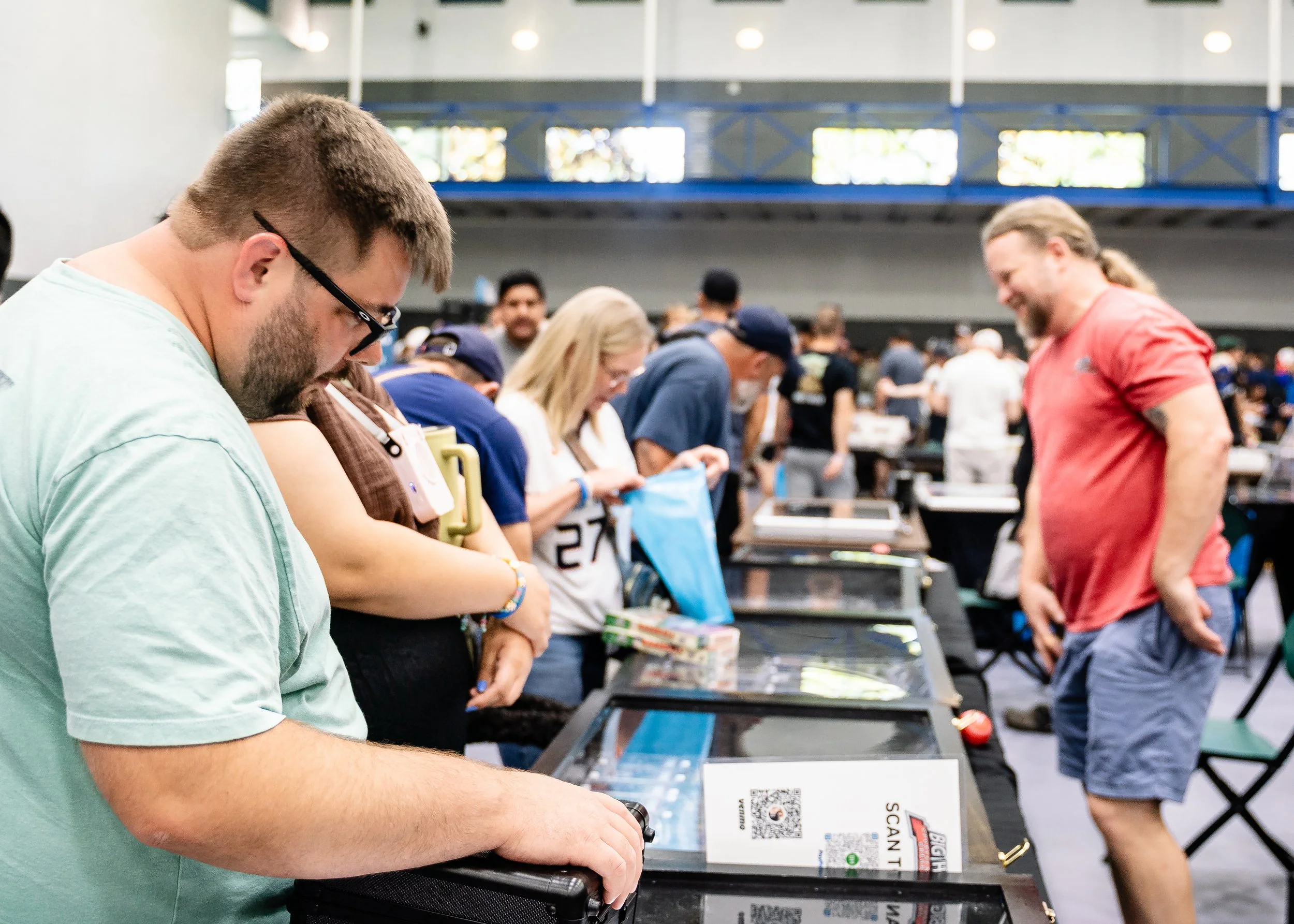 People in a crowded indoor space, possibly at a trade show or convention, looking at tables with various items. A man in a mint green shirt and glasses is in the foreground, while other individuals are in the background, engaging with exhibits or pro