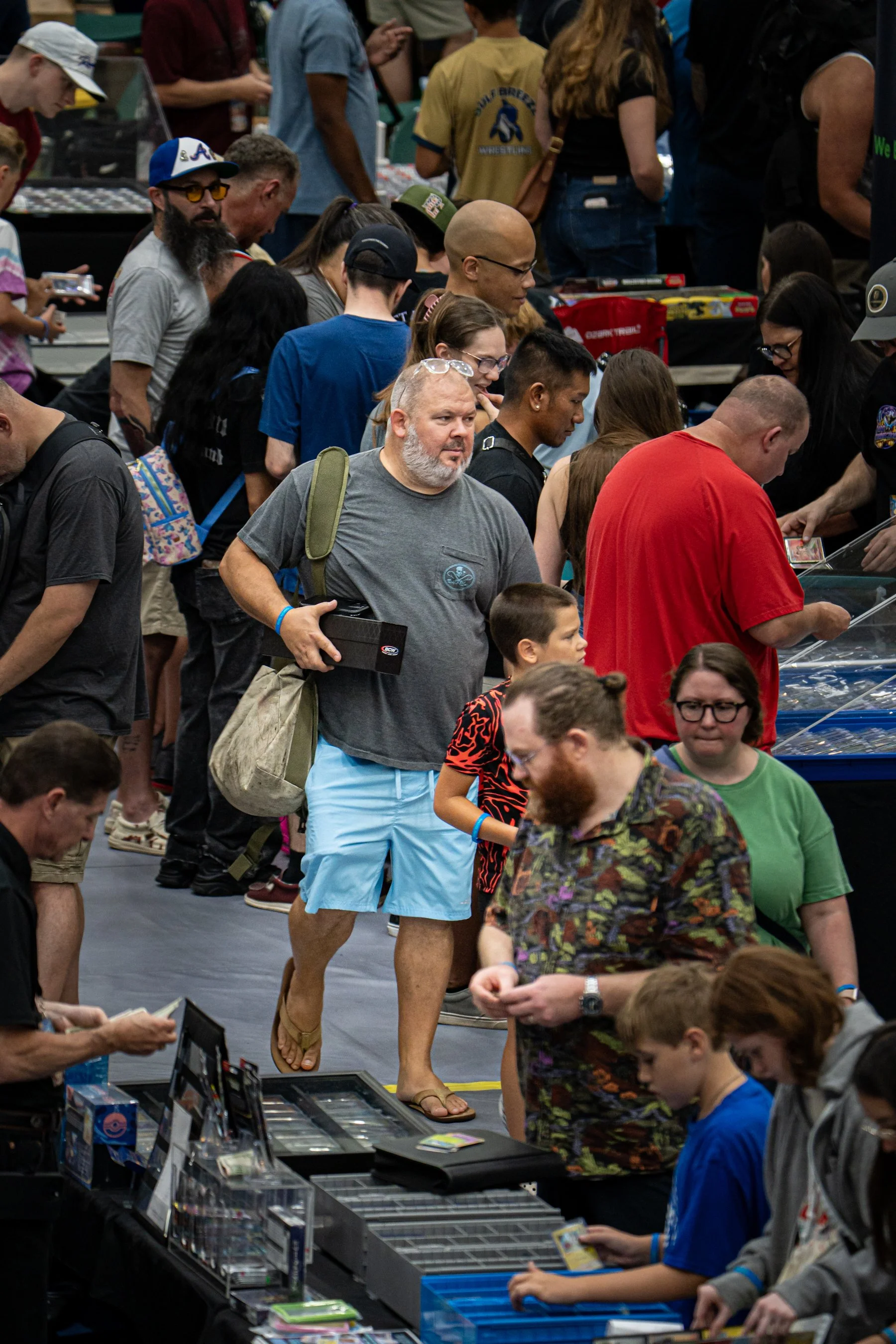 People gathered at a convention or event, browsing tables with various collectibles, games, and merchandise displays.