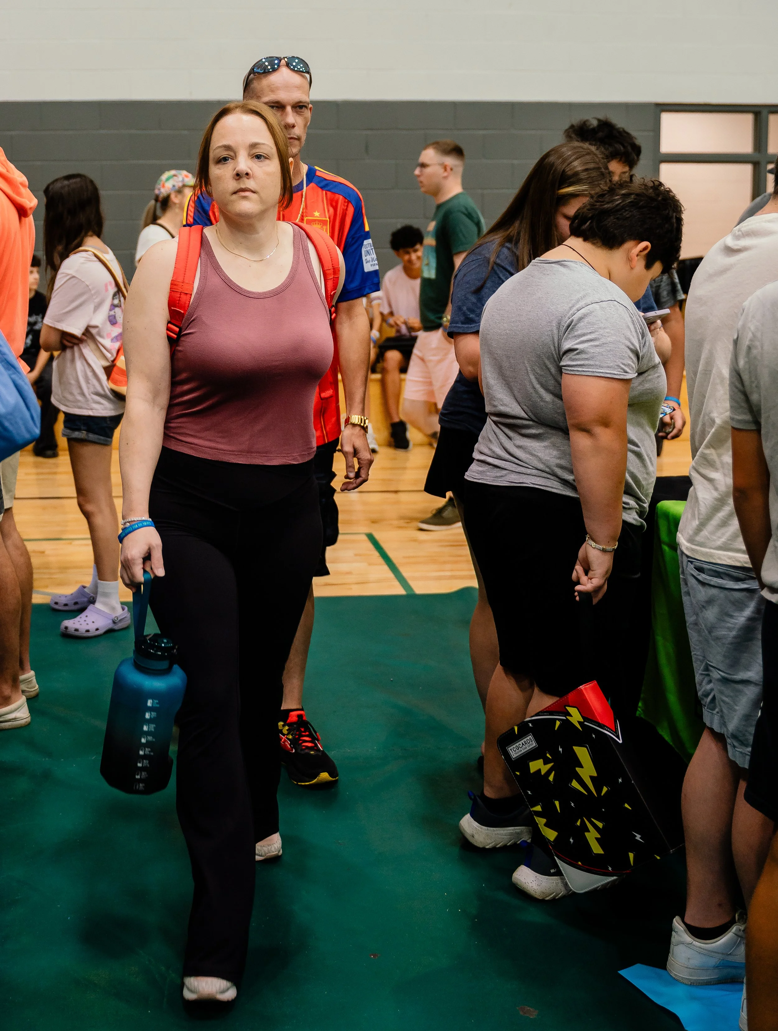 A group of people standing in a gymnasium, some are looking at their phones, a woman with a red backpack and water bottle in hand is in the foreground.