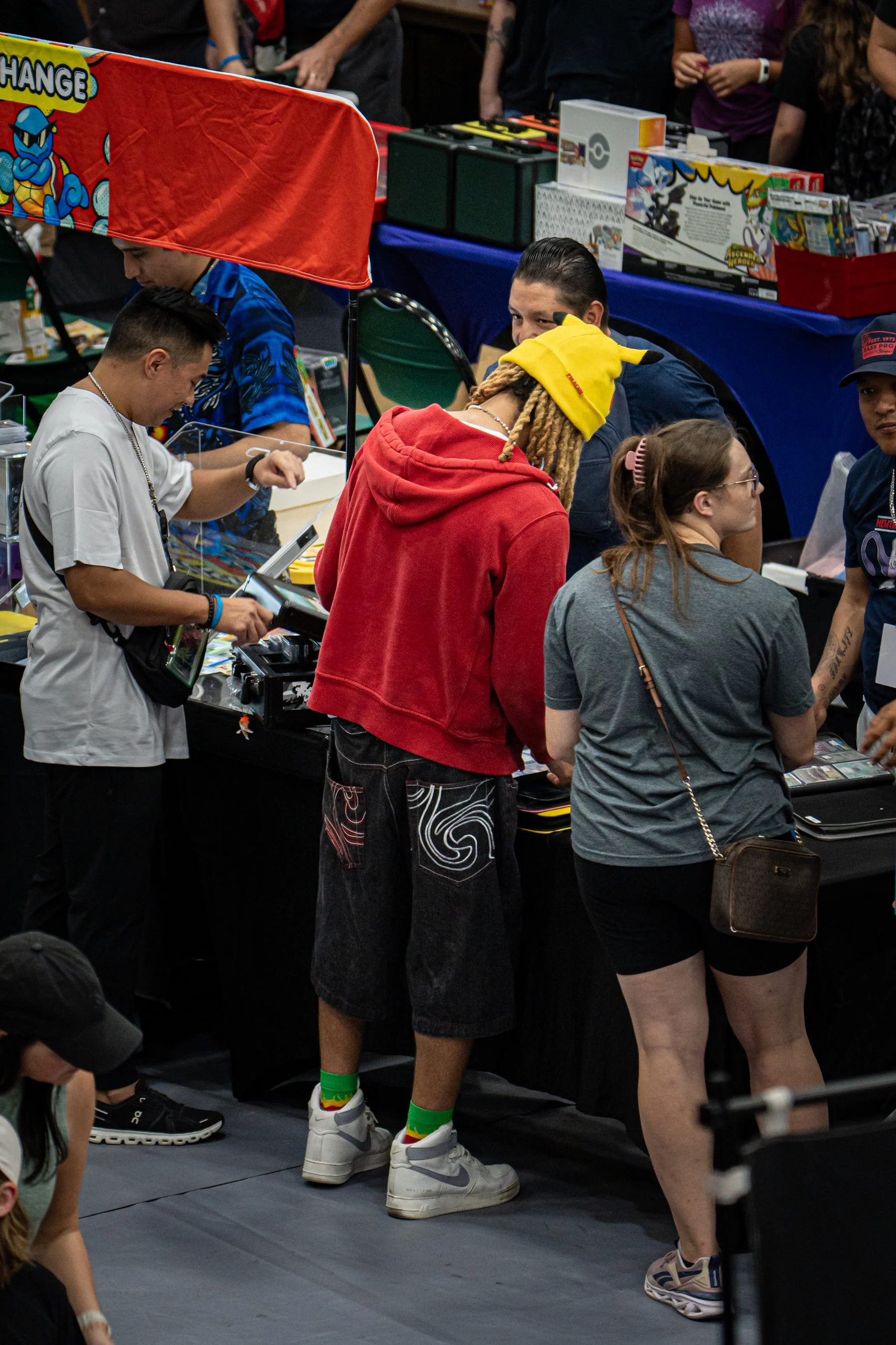 A group of people at a comic or pop culture convention are gathered around a booth with various merchandise. One person is wearing a yellow Pikachu hat, a red hoodie, and colorful socks with sneakers. Others are looking at items on display, and sever