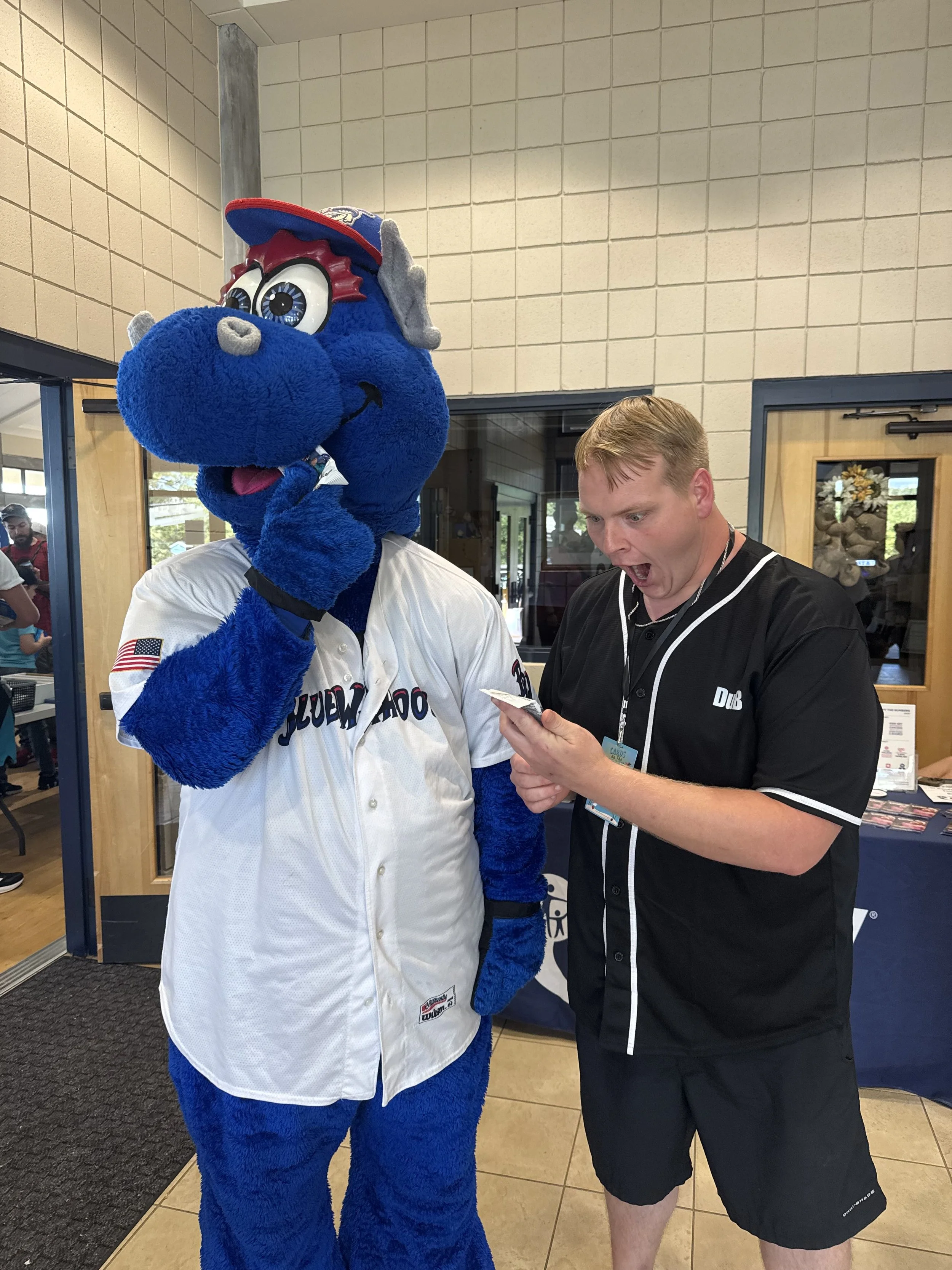 A person in a blue mascot costume of a dragon or dinosaur, wearing a baseball jersey, and an adult male with short blonde hair, wearing a black sports jersey, are indoors near a door with a window and a decorative flower arrangement behind them. The mascot is holding a phone to its ear, and the man looks surprised or excited as he looks at the phone.