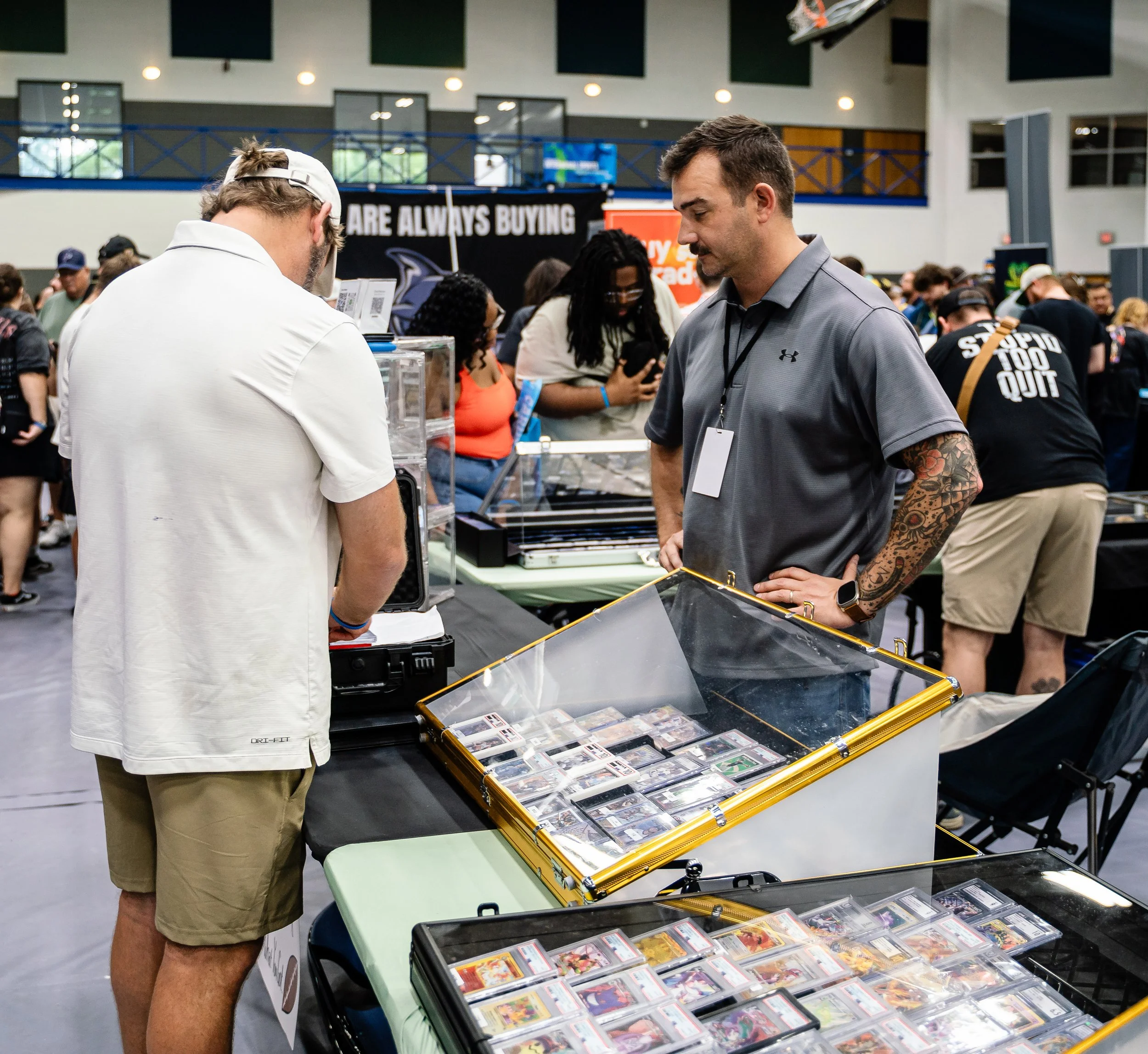 Two men looking at trading cards and memorabilia at a convention or trade show, with a table displaying cards and people browsing in the background.
