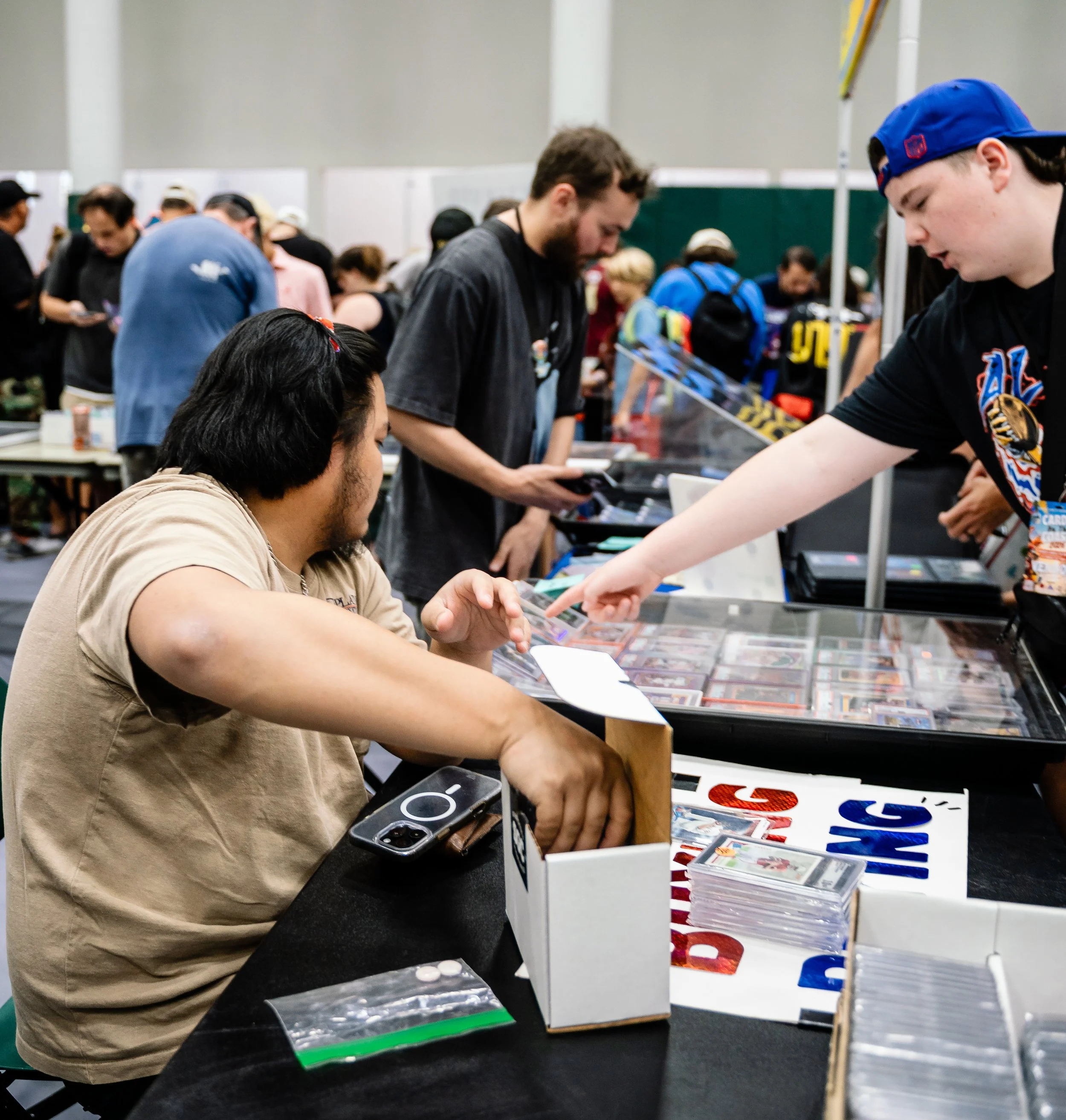 People at a convention or trading event examining and selling trading cards, with stacks of cards displayed on tables and attendees browsing and chatting.