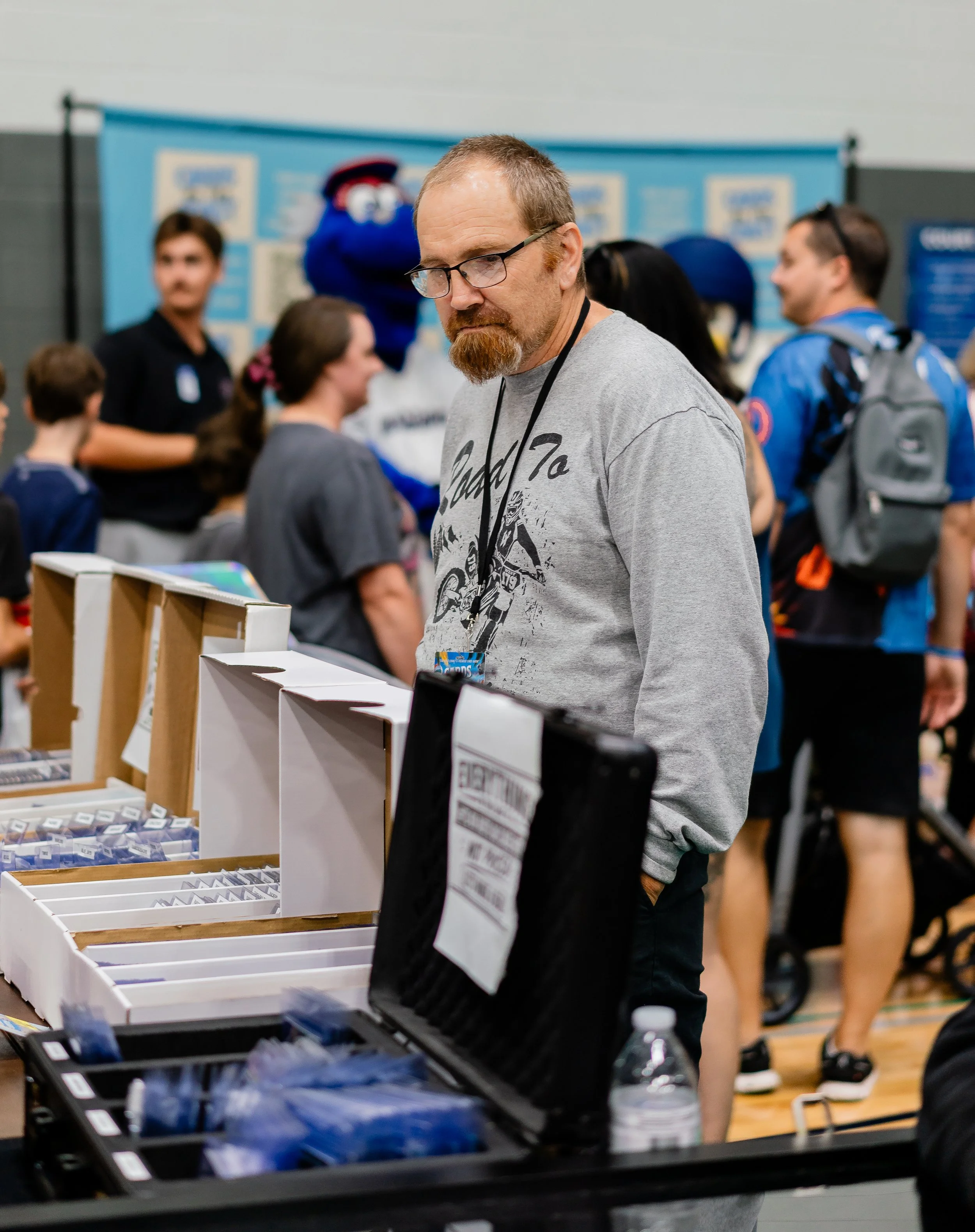 A man with glasses and a beard stands at a table with various small items, in a busy indoor setting with people and a mascot in the background.