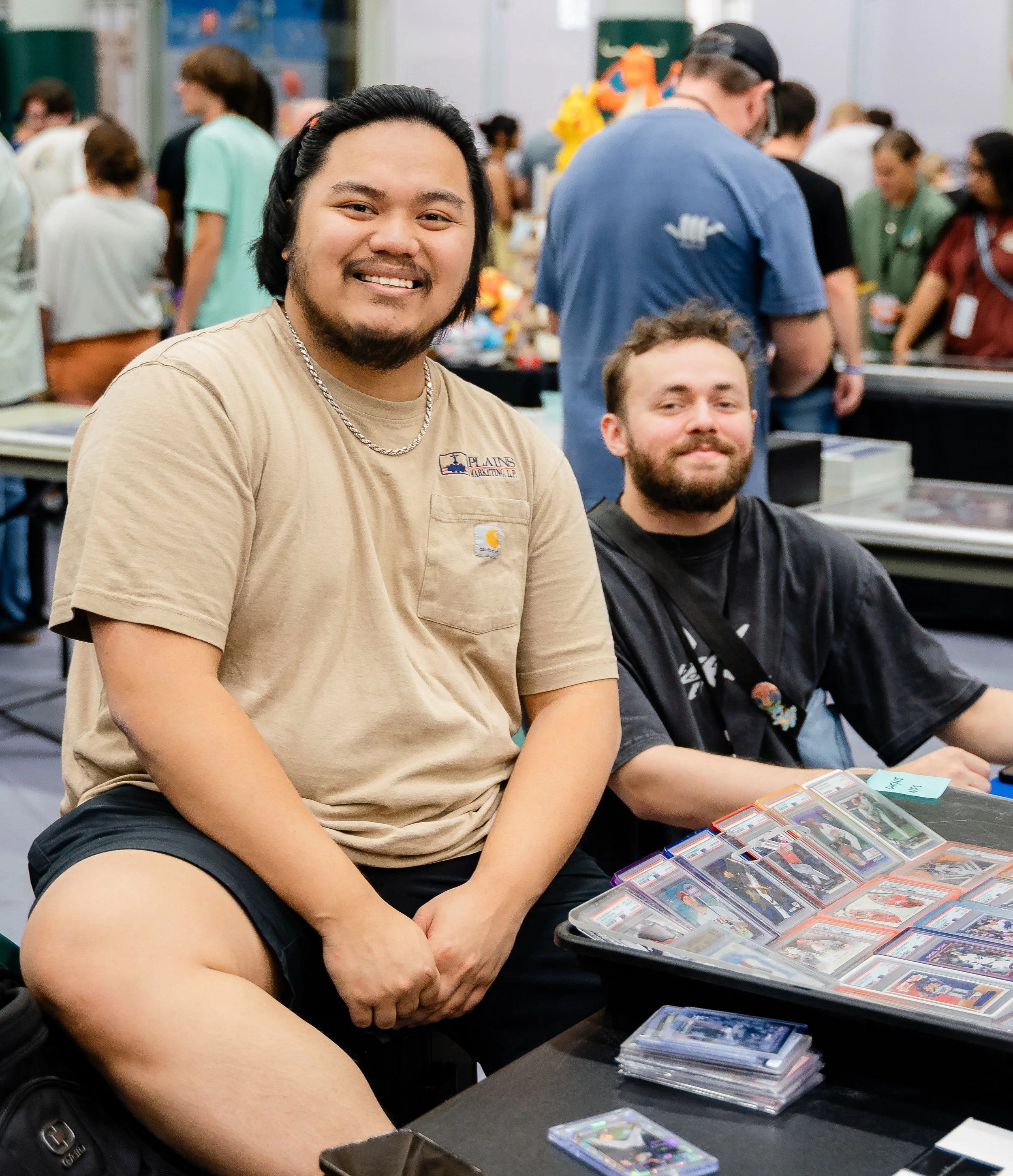 Two young men sitting at a table during a trading card event, with trading card collections in front of them. One is smiling and wearing a beige T-shirt, while the other, with a beard and wearing a black T-shirt, is looking at the cards. The backgrou