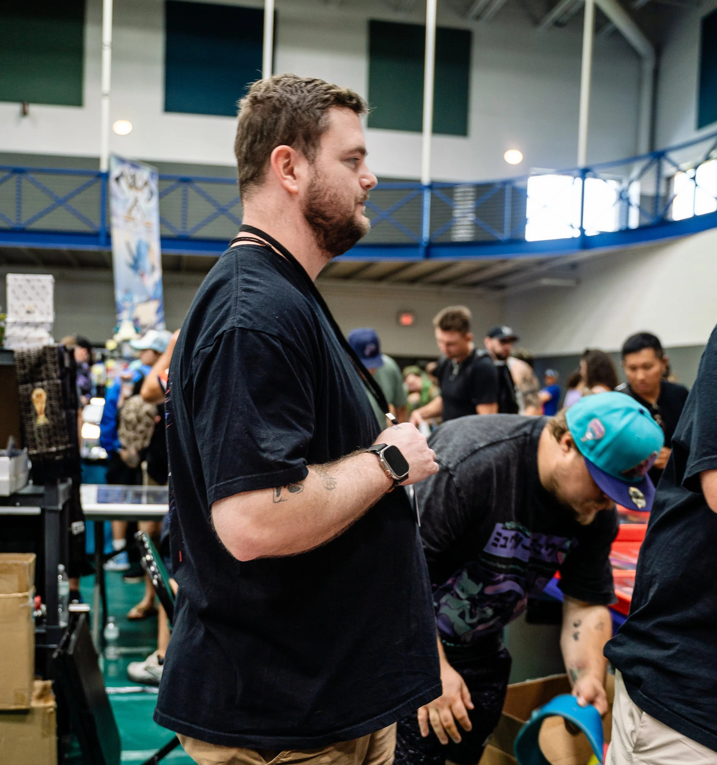 A man with short hair and a beard, wearing a black t-shirt, is standing indoors at a crowded event. He is holding a smartphone and appears focused on it. There are many people around, some with hats, in a space that looks like a convention or exhibit