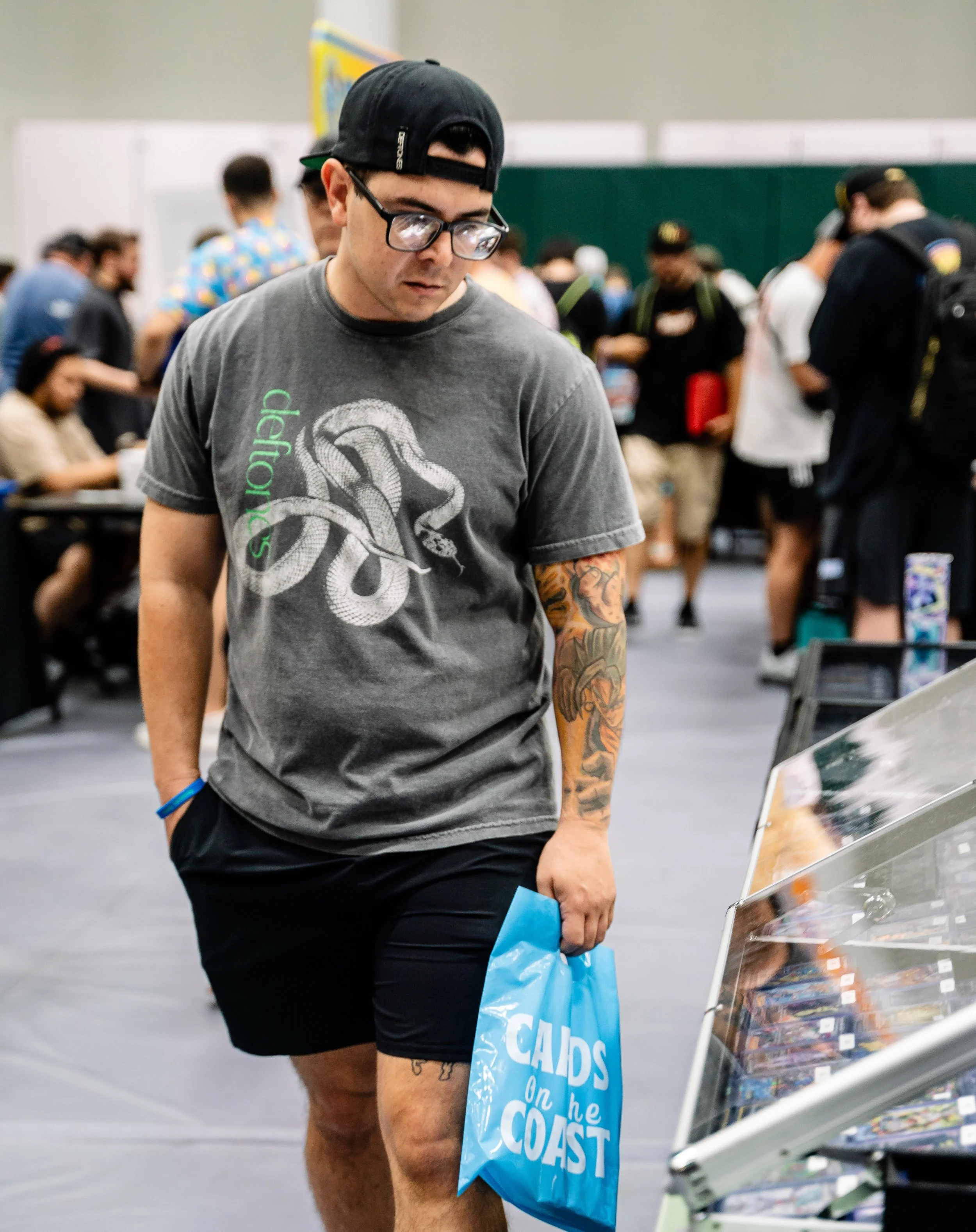 Young man with glasses and a backwards cap, wearing a gray T-shirt with a snake graphic, looking at a rack of DVDs or video games.