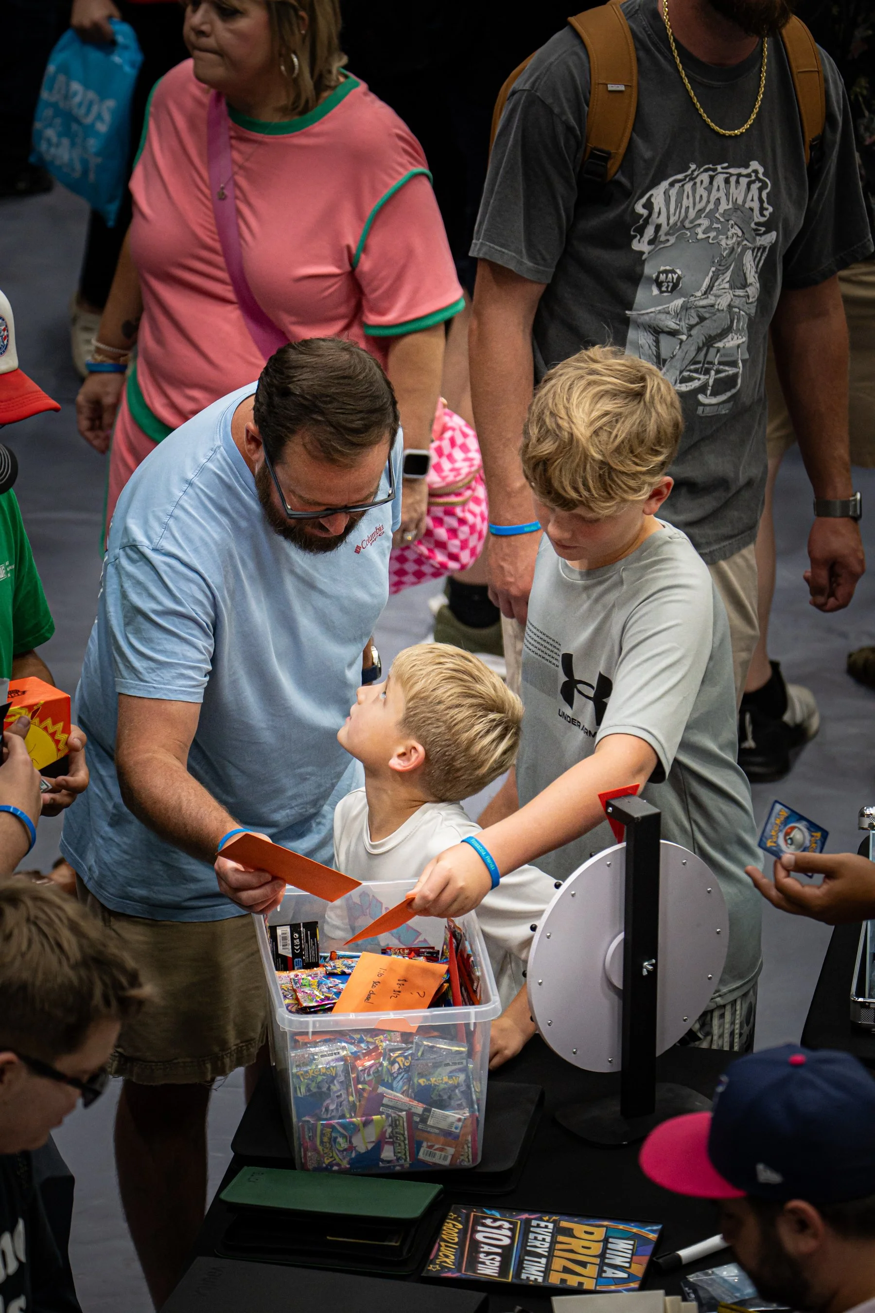 People gathered around a table at a convention or event, looking at items in a plastic container filled with Pokémon trading cards. The scene includes children and adults, some holding cards or involved in the activity, with merchandise and promotion