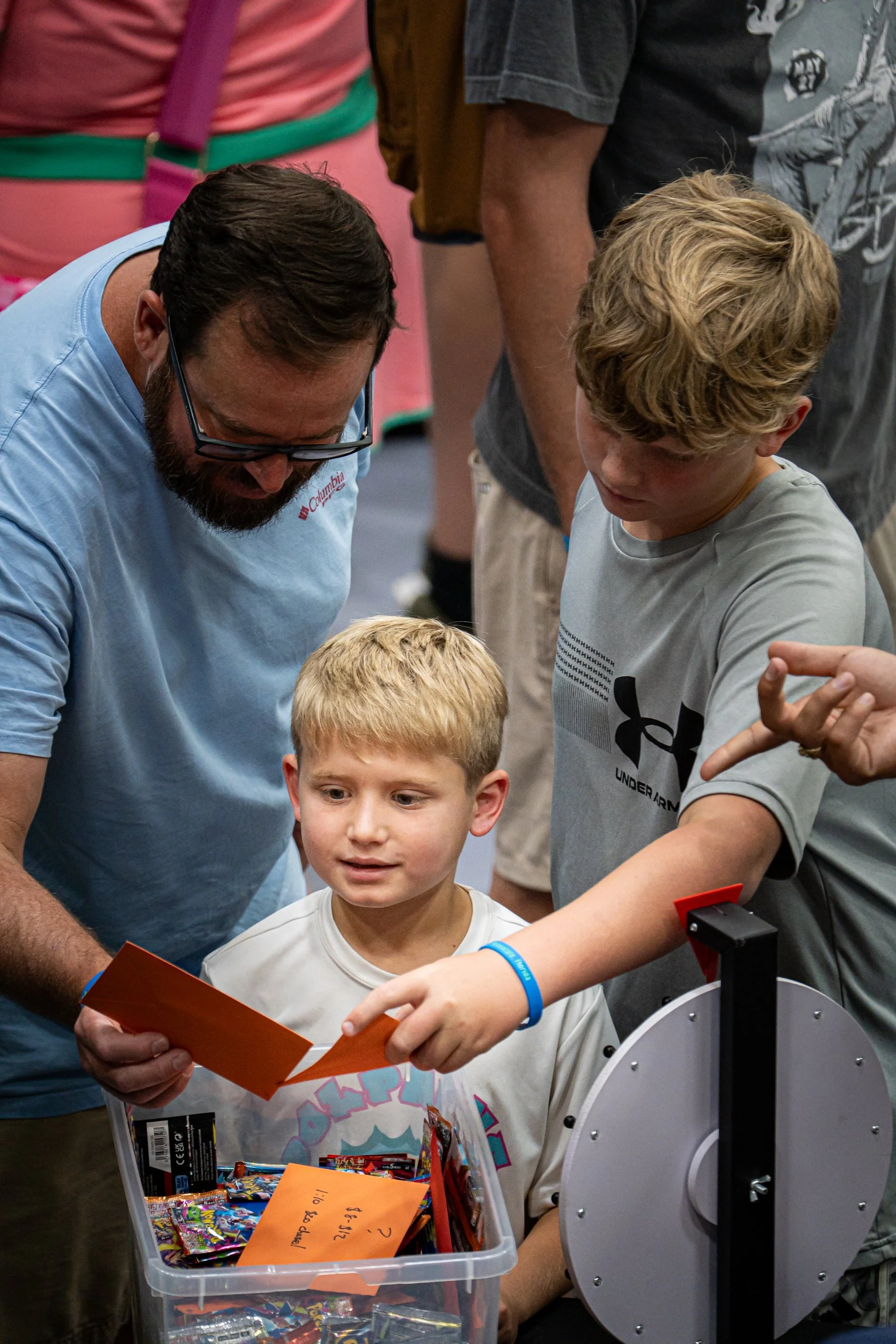 A young boy is grabbing candies from a donation box, with two adult men looking on. The scene appears to be at a public event or fundraiser.