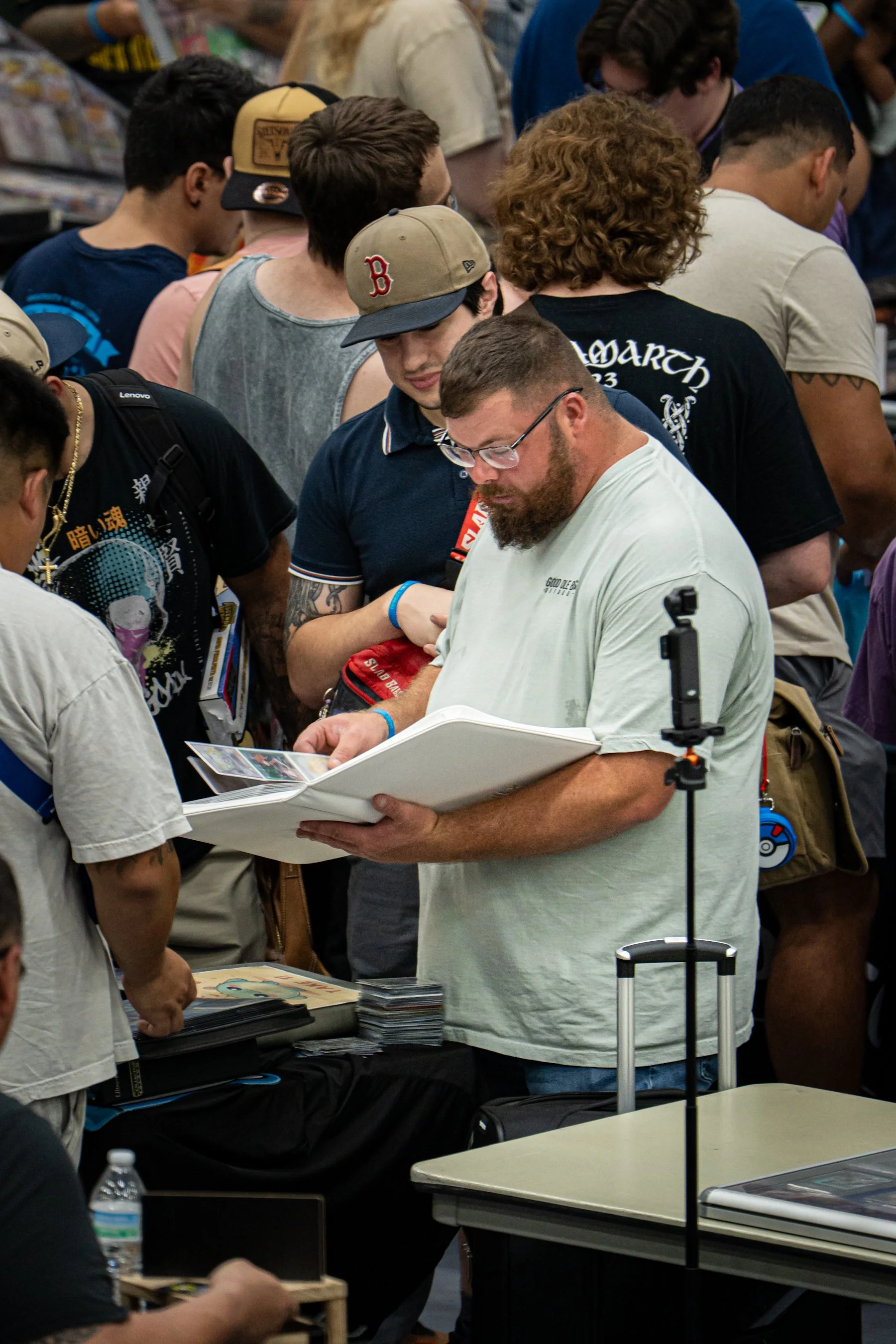 A crowd of people standing at a table and looking at displays or signing materials at an event, possibly a convention or signing event.