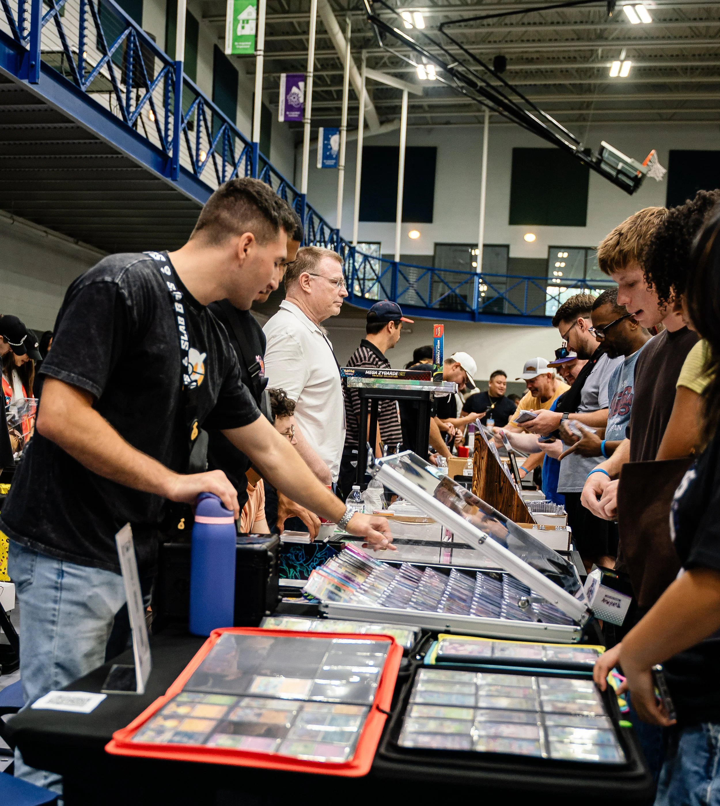 People browsing table of toys and collectibles at convention or trade show in large indoor venue with high ceiling and banners.