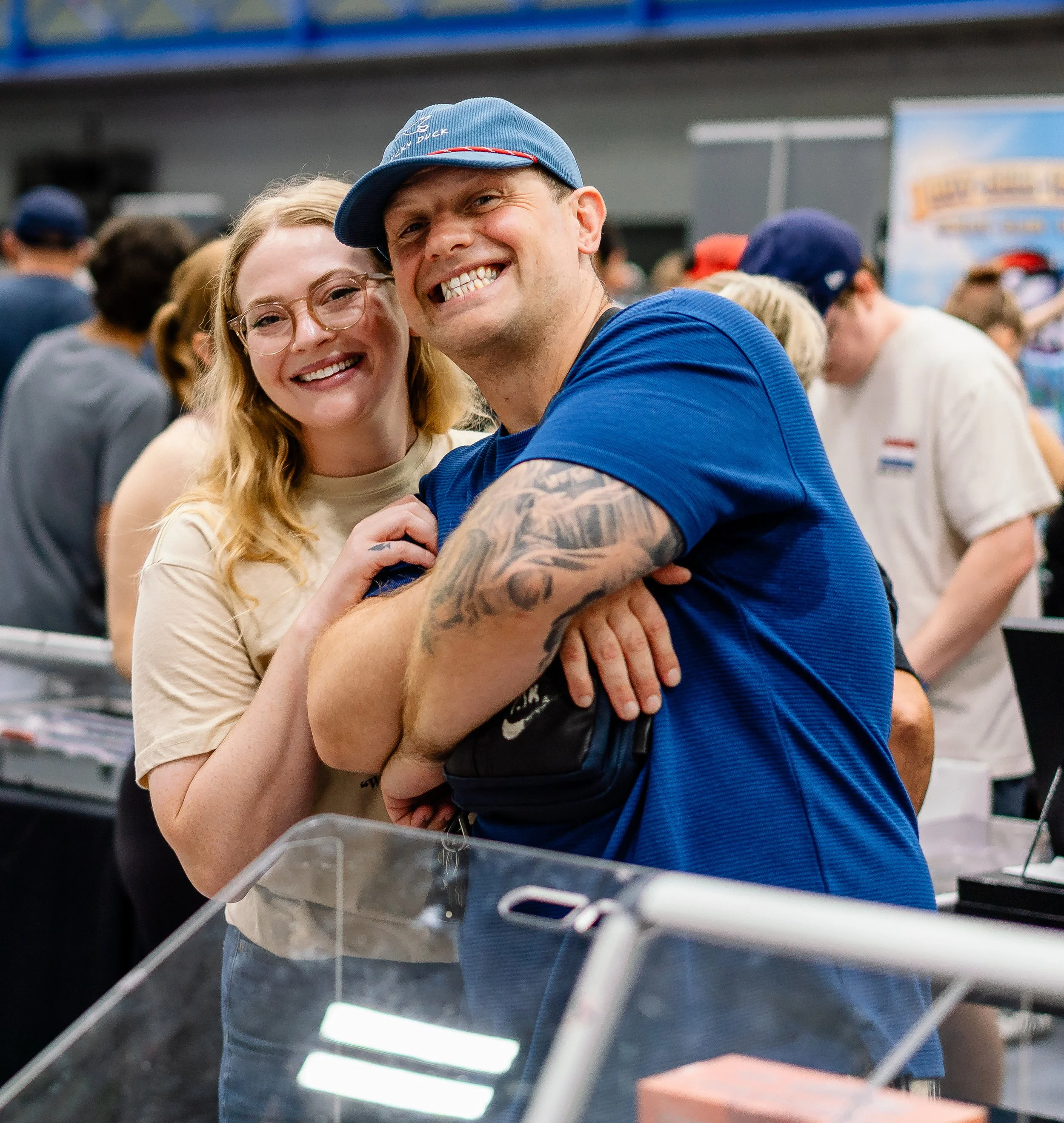 A smiling couple with a man wearing a blue cap and a woman with glasses, both looking at the camera at what appears to be a store.