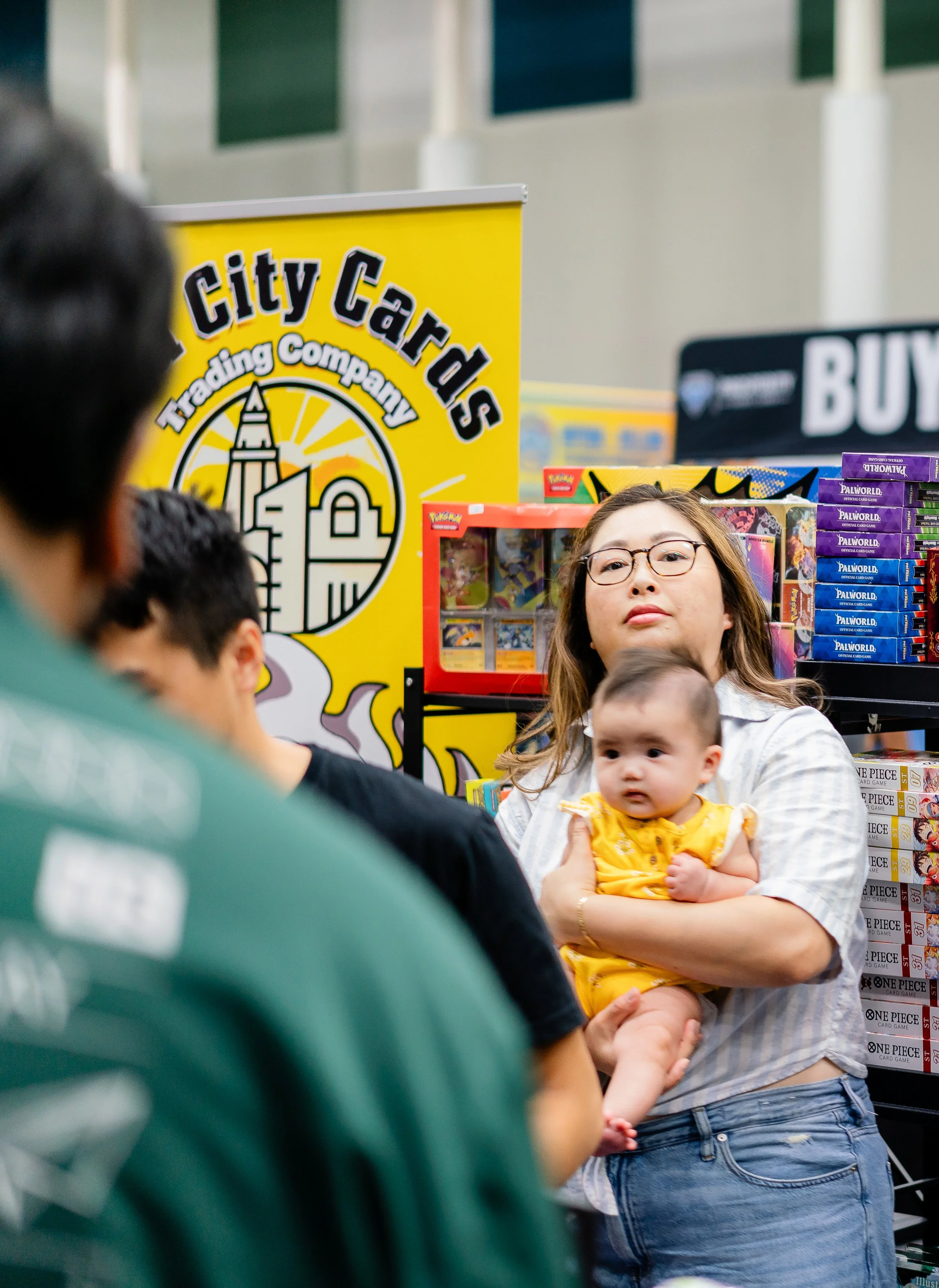 A woman holding a baby in front of a display of Pokémon cards at a store, with a yellow City Cards Tradeing Company sign visible in the background.