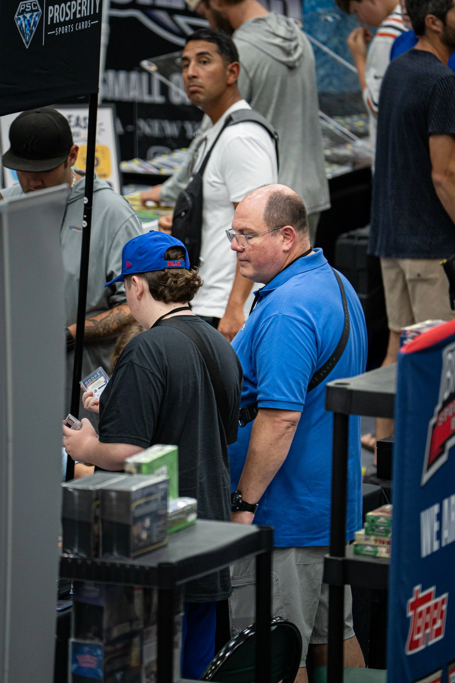 People at a booth or store buying sports cards, with a banner displaying 'Prosperity Sports Cards' and various boxed items on display.