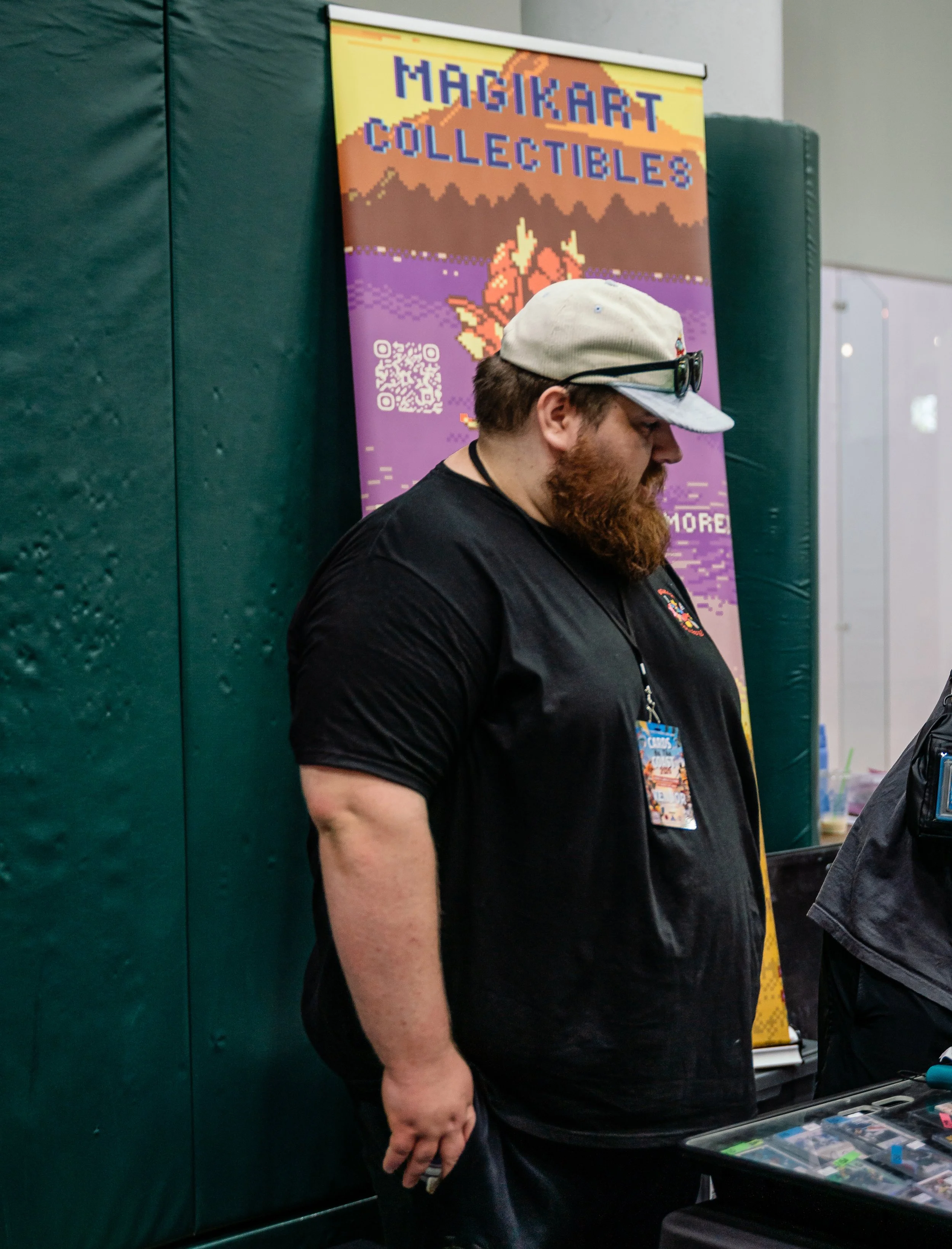 A man with a beard, wearing a light-colored cap and sunglasses on top, is looking down at items on a table. Behind him is a colorful poster that reads 'Magikart Collectibles' with a QR code and a pixel art graphic. The setting appears to be an indoor