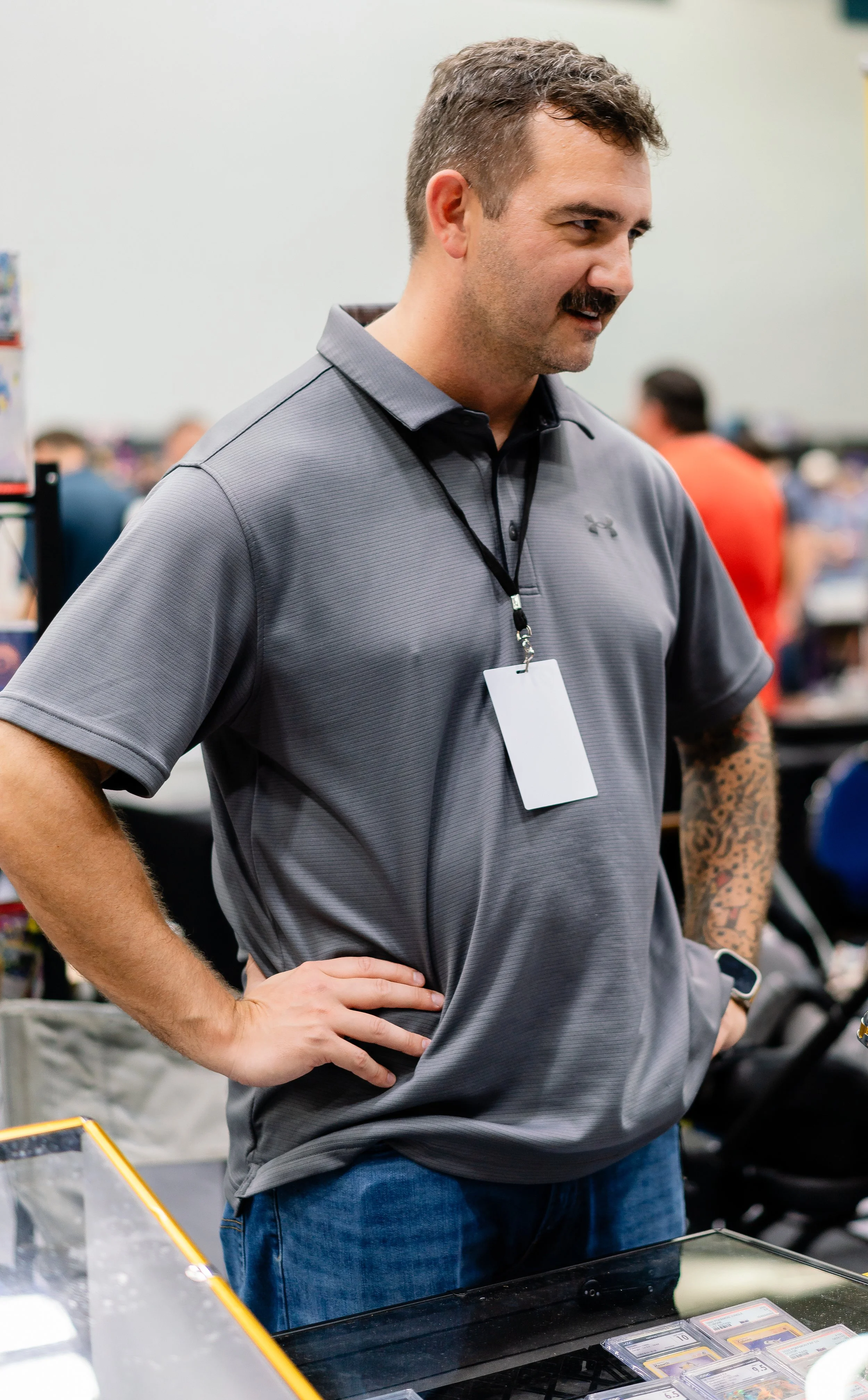 A man with a mustache, wearing a gray polo shirt and blue jeans, stands with one hand on his hip at a display case with collectible cards, in a busy indoor setting.
