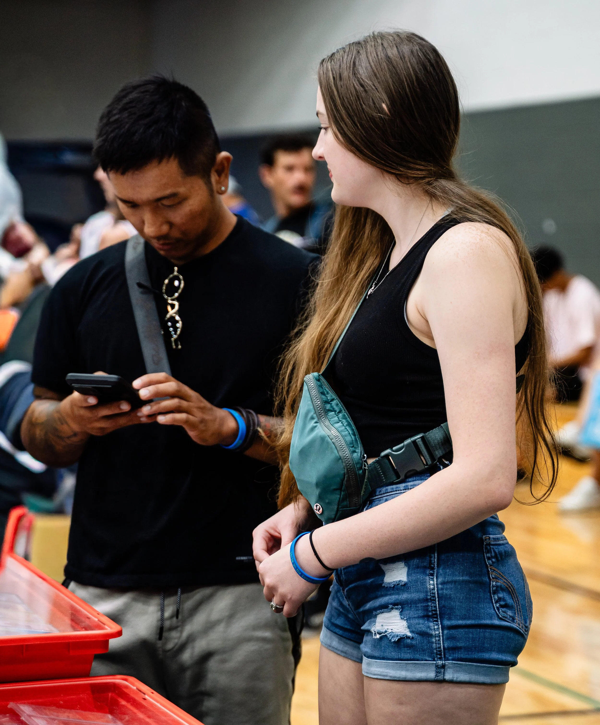 Two women standing side by side at a table, one looking at her phone and the other smiling, in a social or event setting with people in the background.