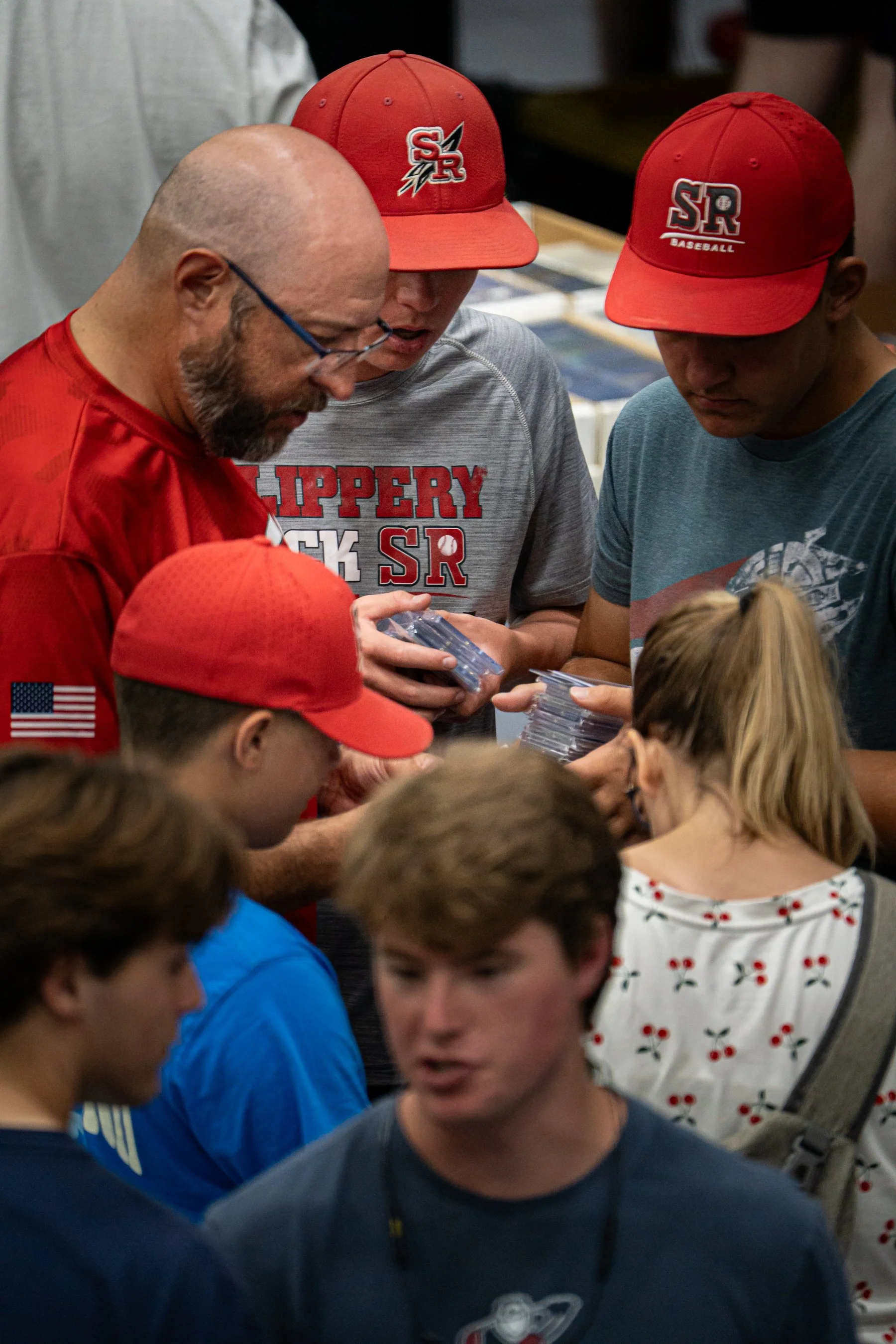A group of people, including children and adults, gathered around looking at items in their hands. Some are wearing red caps with sports logos, and a few adults are wearing sports or casual attire.