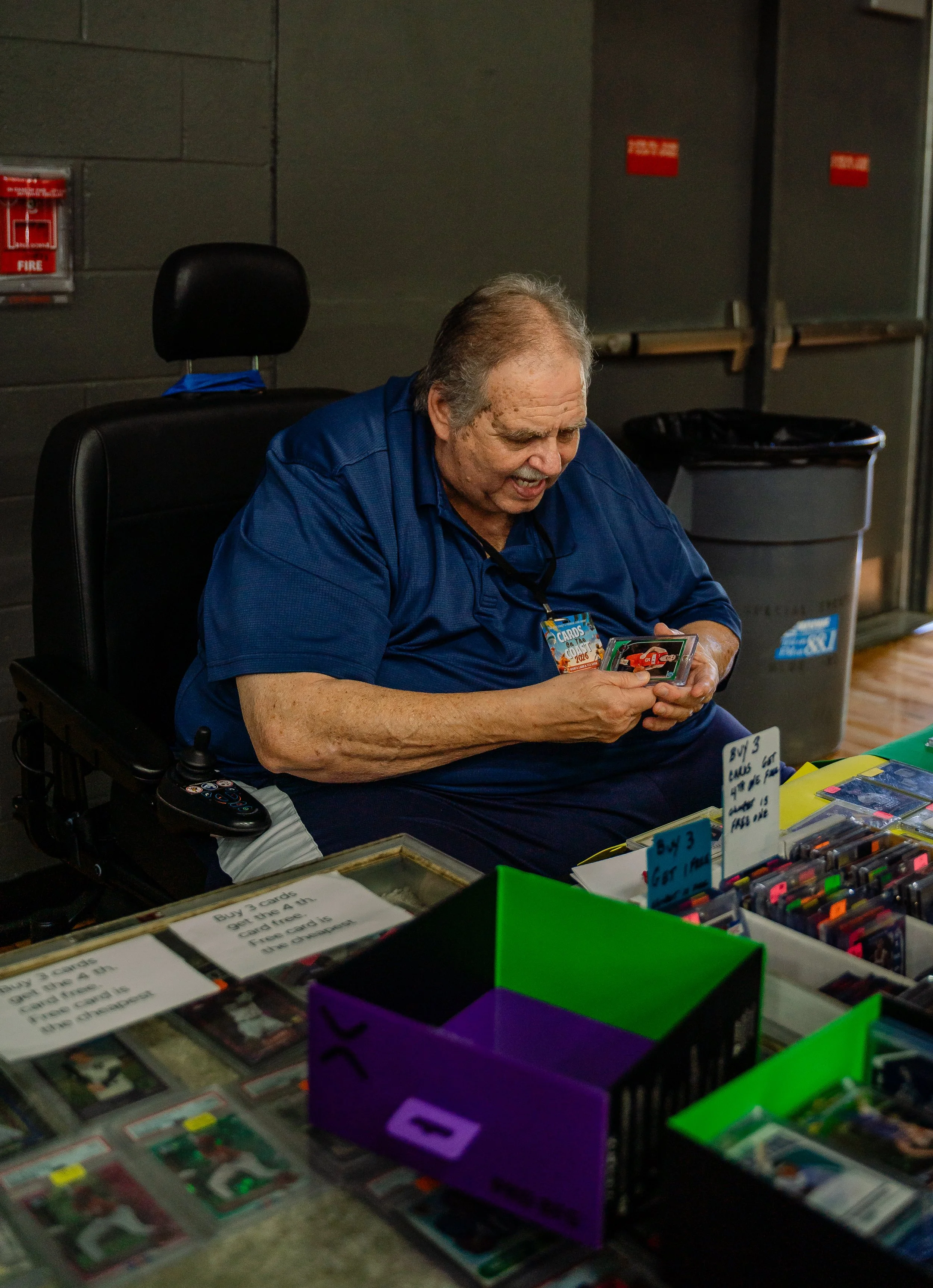 An elderly man with gray hair and a mustache, wearing a blue shirt, is sitting at a table with various trading cards and collectible cards. He is smiling and looking at a card he is holding. There are signs and containers with more cards on the table