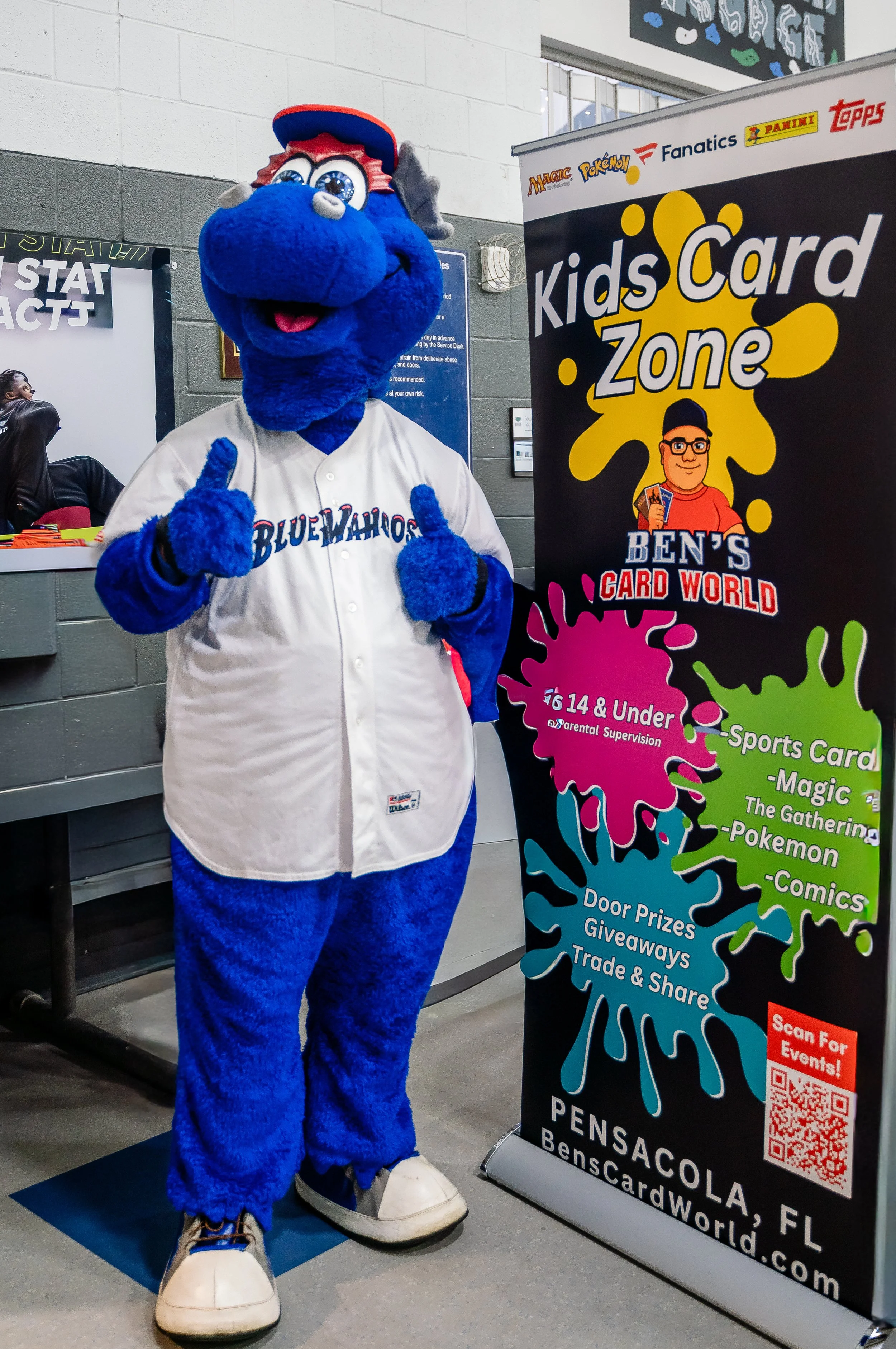 Blue mascot dressed in a White baseball jersey with 'Blue Wahoos' written on it, standing in front of a Kids Card Zone banner at an indoor event.