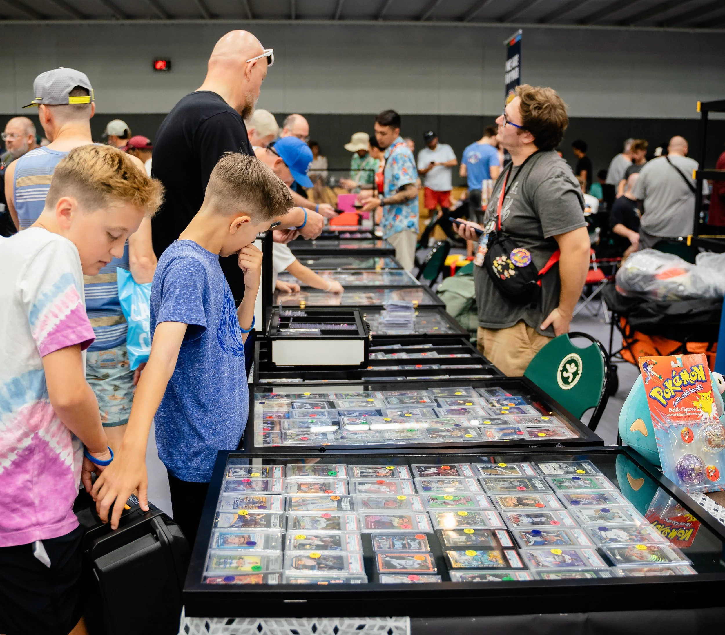 Multiple people browsing and purchasing items at a table filled with trading cards and Pokémon merchandise at an indoor convention or event.