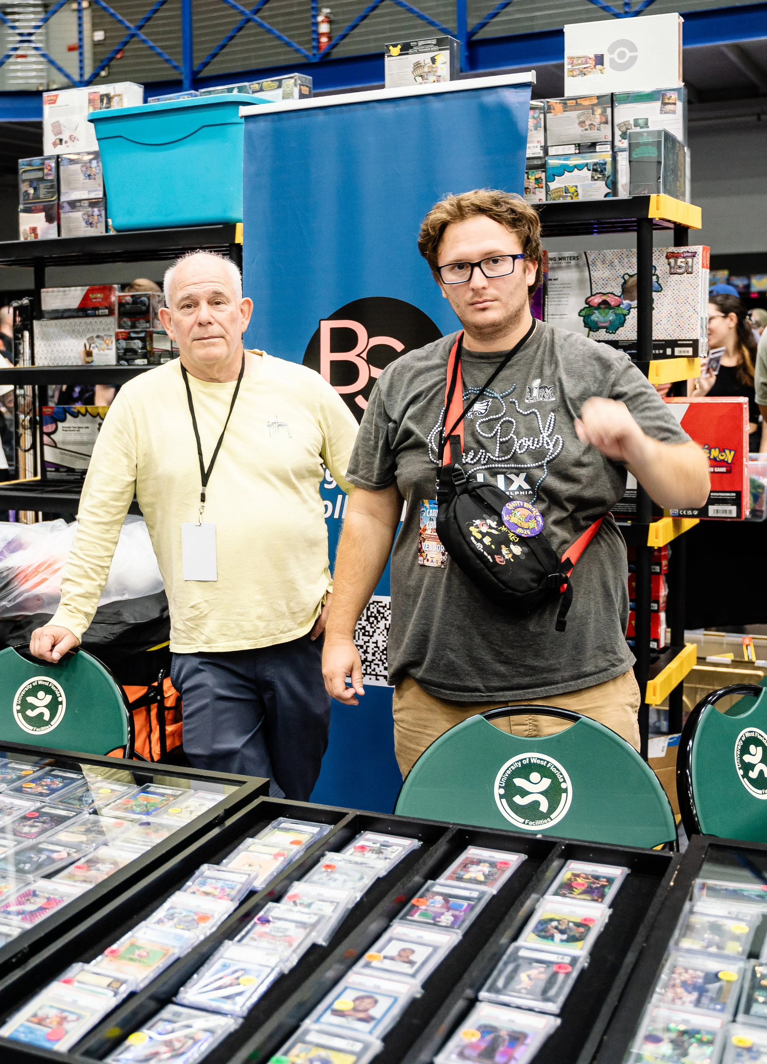 Two men standing behind tables with collectible trading cards in plastic cases at a convention or trade show, with shelves of boxed collectibles in the background.