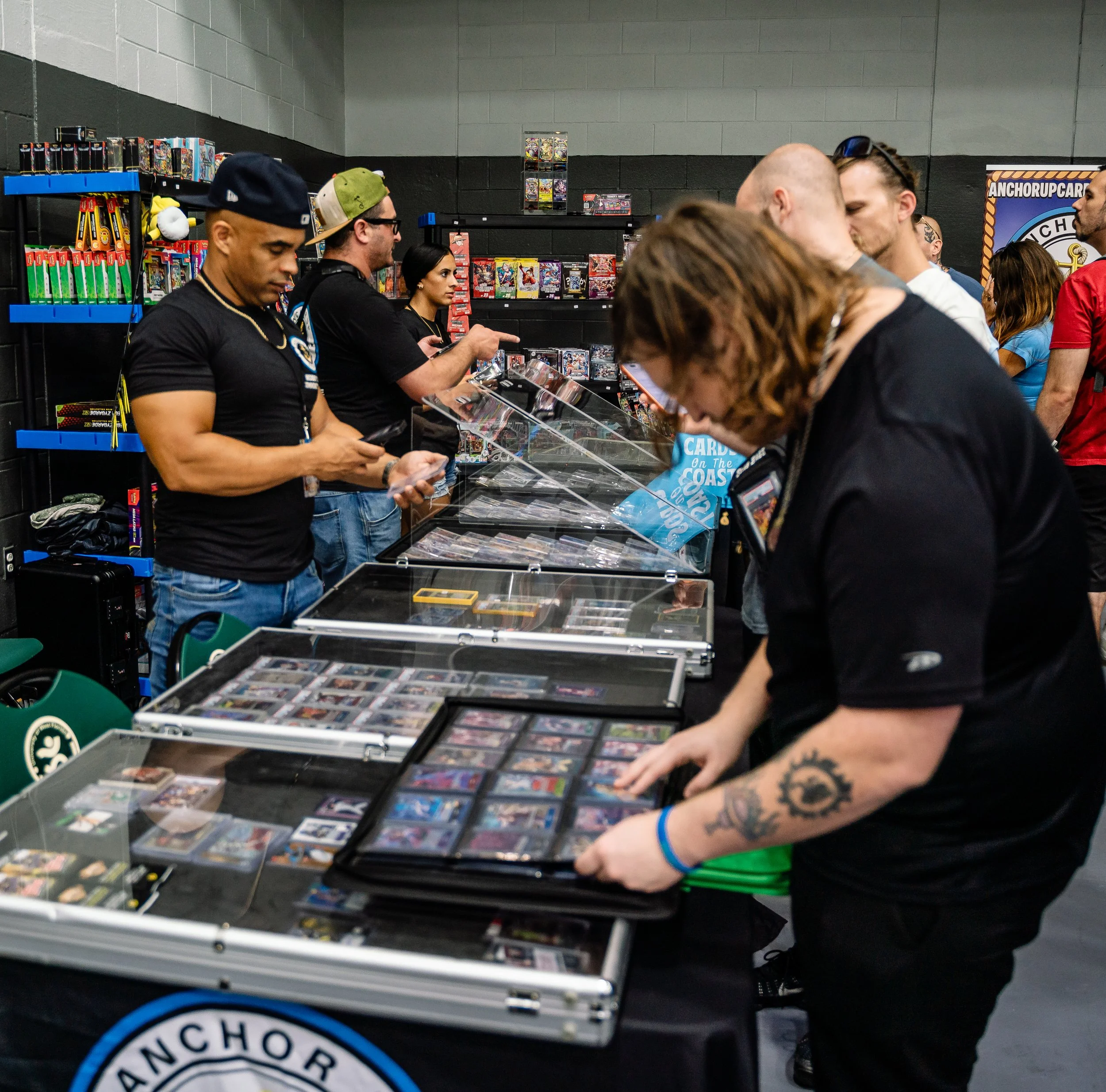 Several people shopping for trading cards at a table filled with card packs and cards, shelves with more card packs in the background, inside a store with black and gray walls.