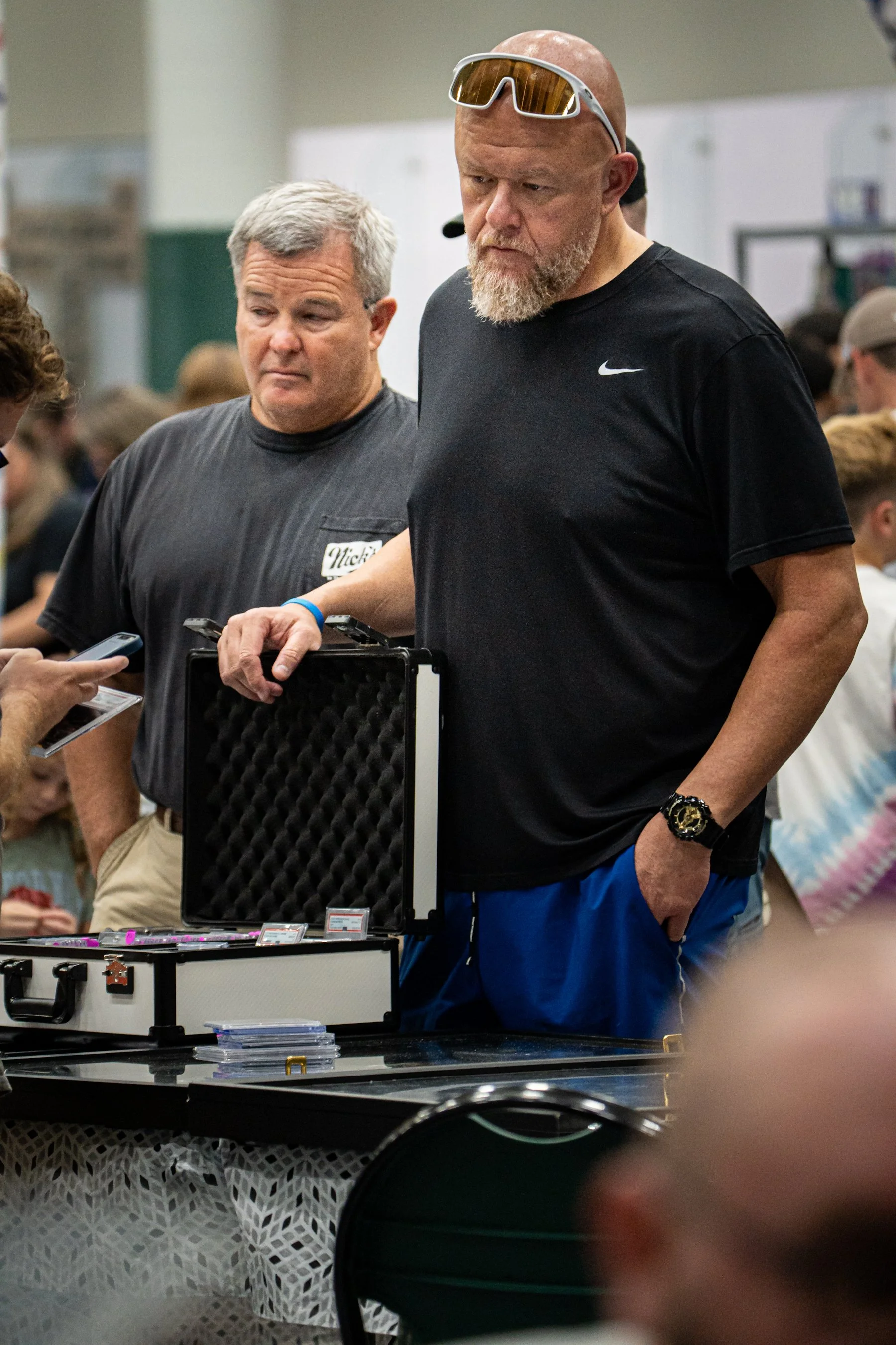 Two men at a table examining trading cards or collectible items, with one man holding a black case and a woman nearby showing her phone, in what looks like a marketplace or convention setting.