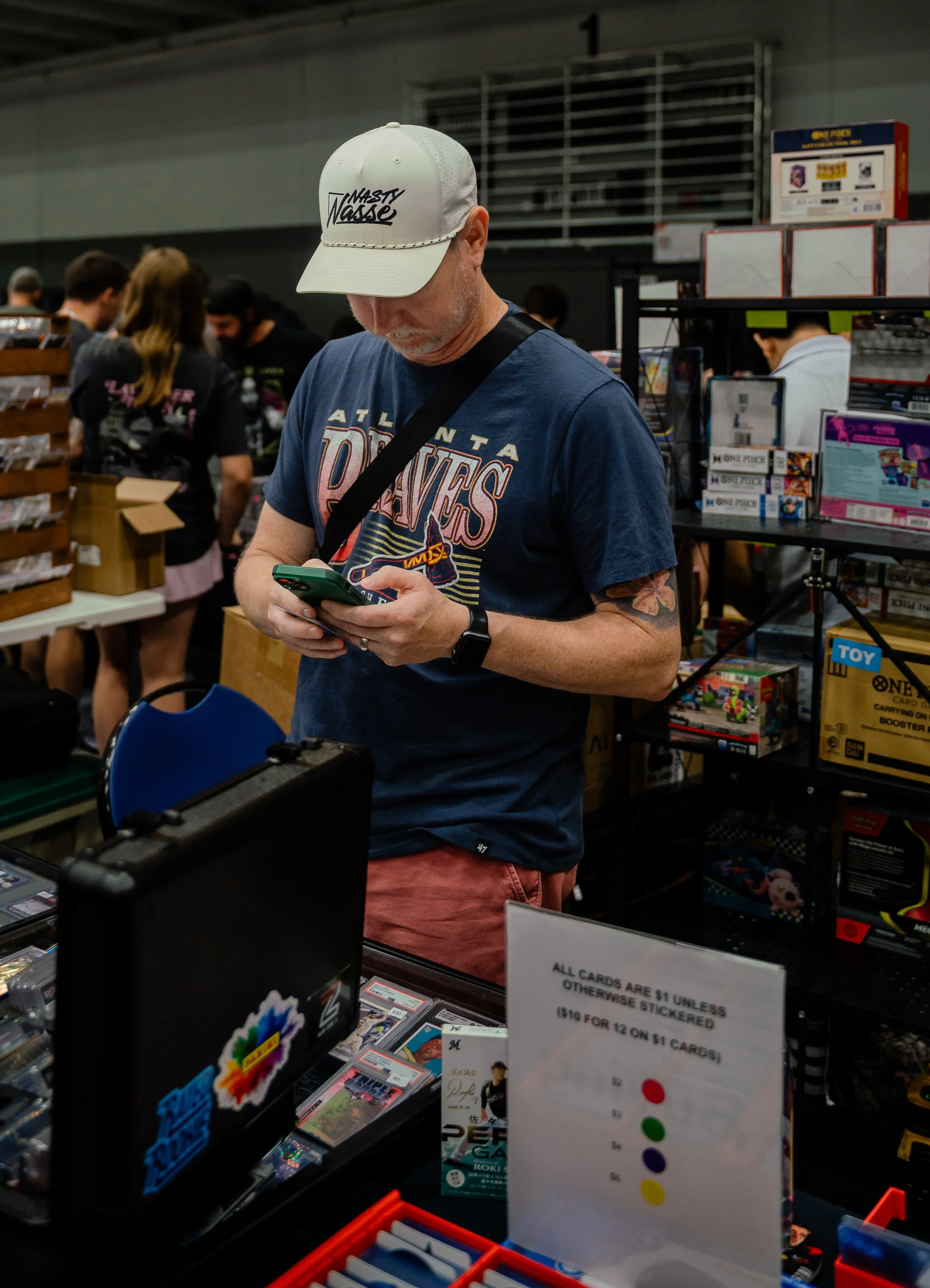 A man wearing a white cap with 'Nasty Nasse' written on it, a blue T-shirt, and red shorts is looking at his phone at a table in a gaming or trading card store. Behind him are shelves with various card and toy boxes, and other people shopping.