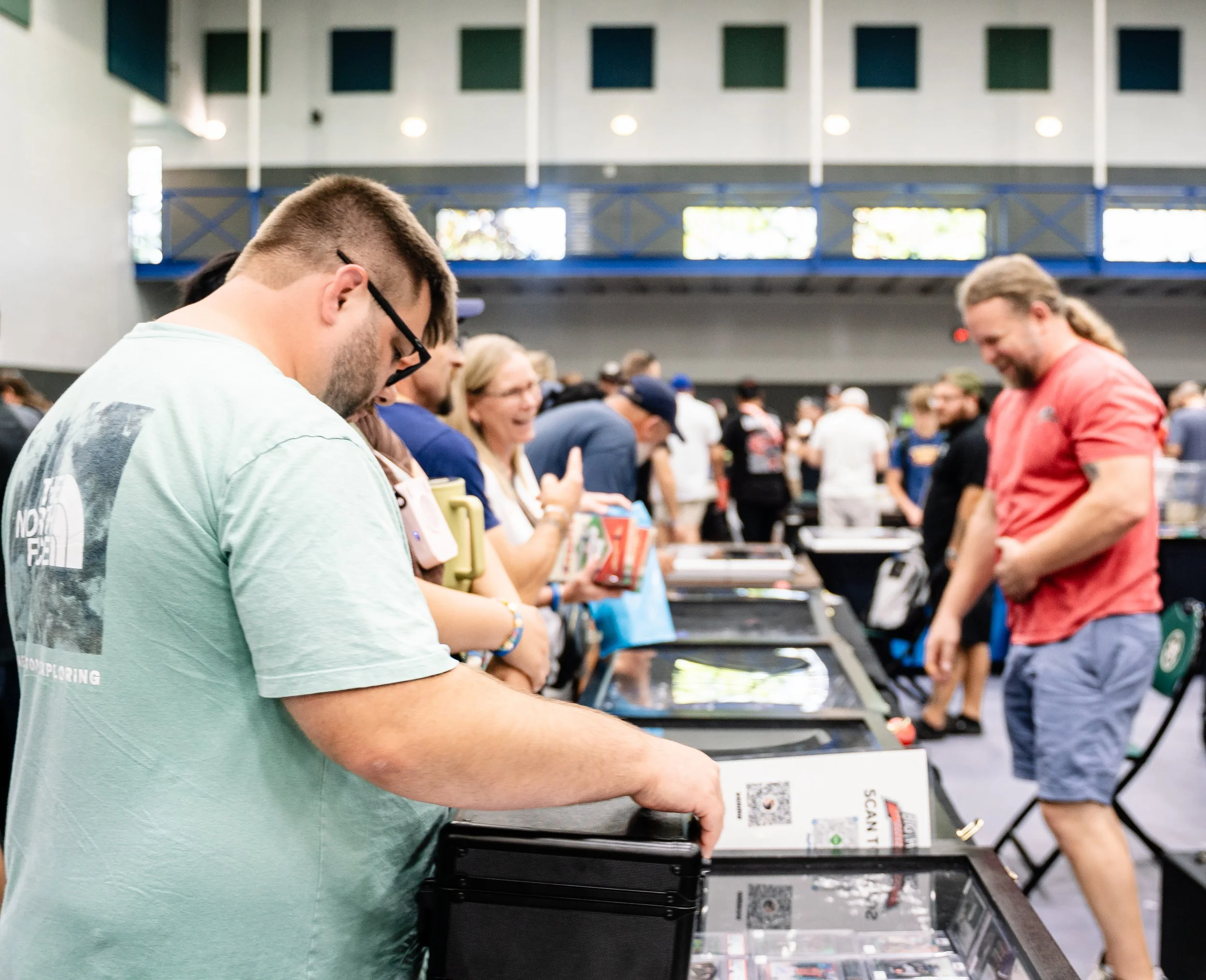 People standing in line at a registration or check-in table inside a large indoor venue, possibly a conference or event.