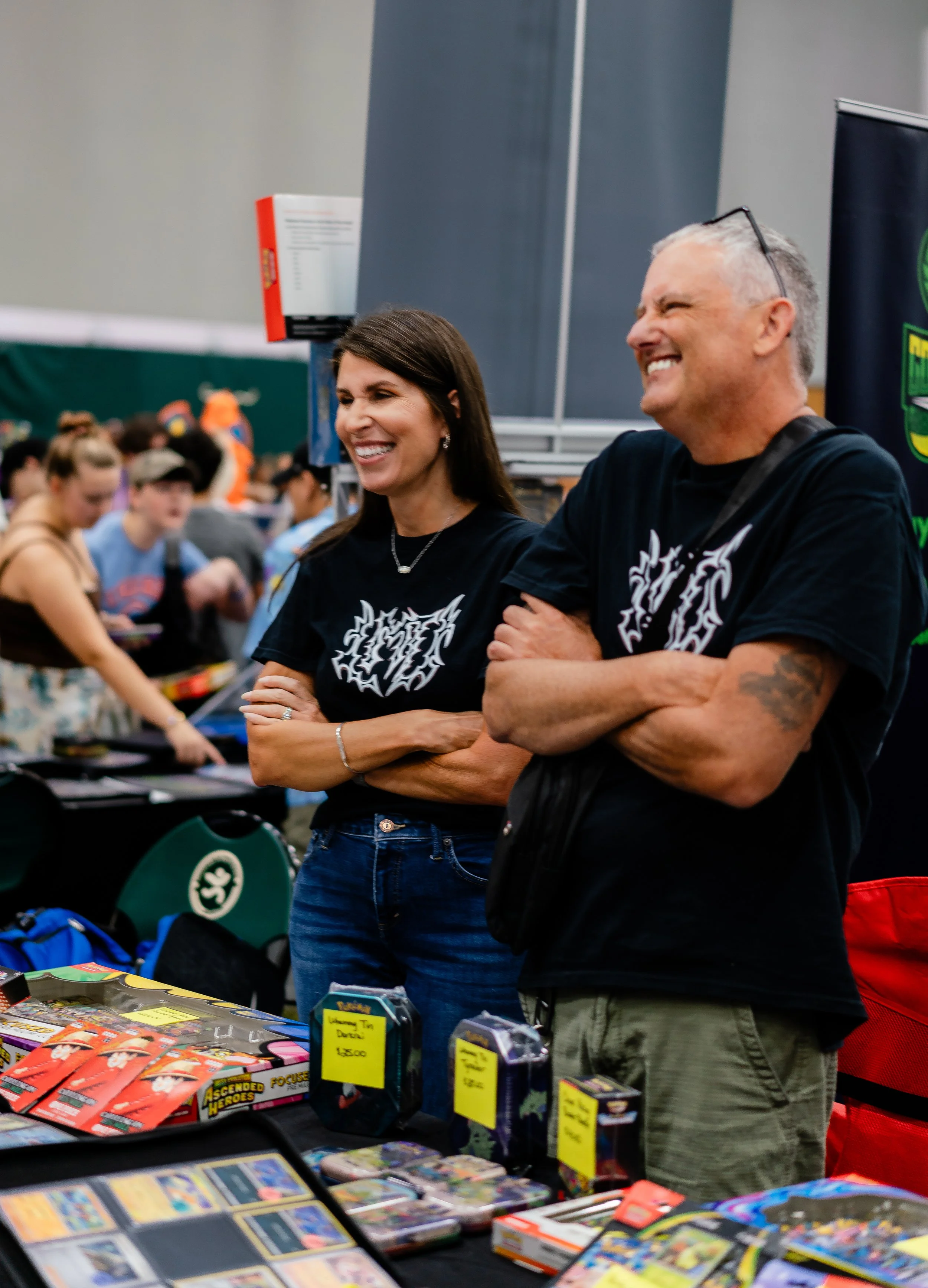 Two smiling people browsing at a table full of comic books and trading cards at a convention or market, with a crowded background and vendor booths.