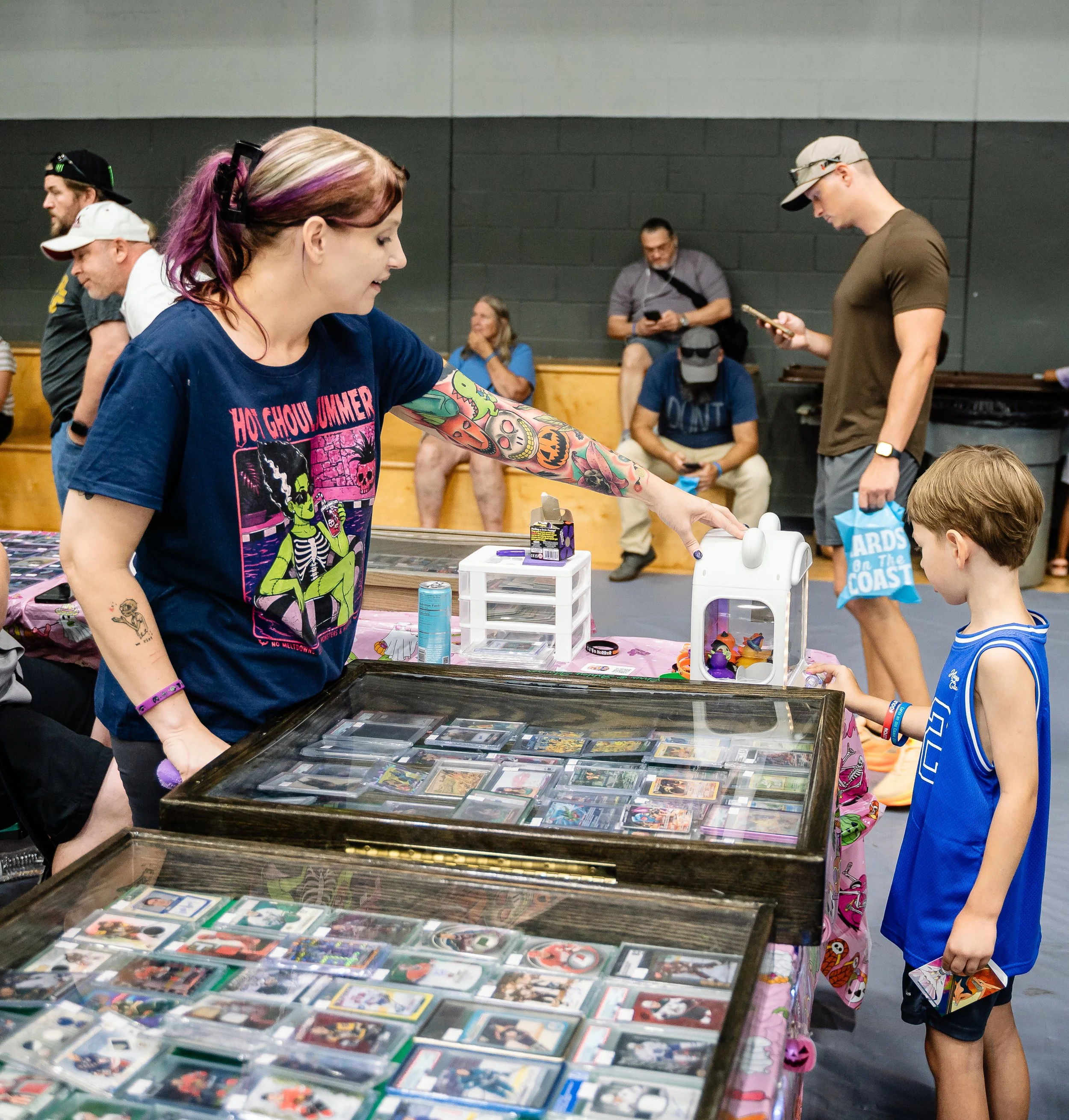 People browsing a table of trading cards at an indoor event, with various spectators in the background.