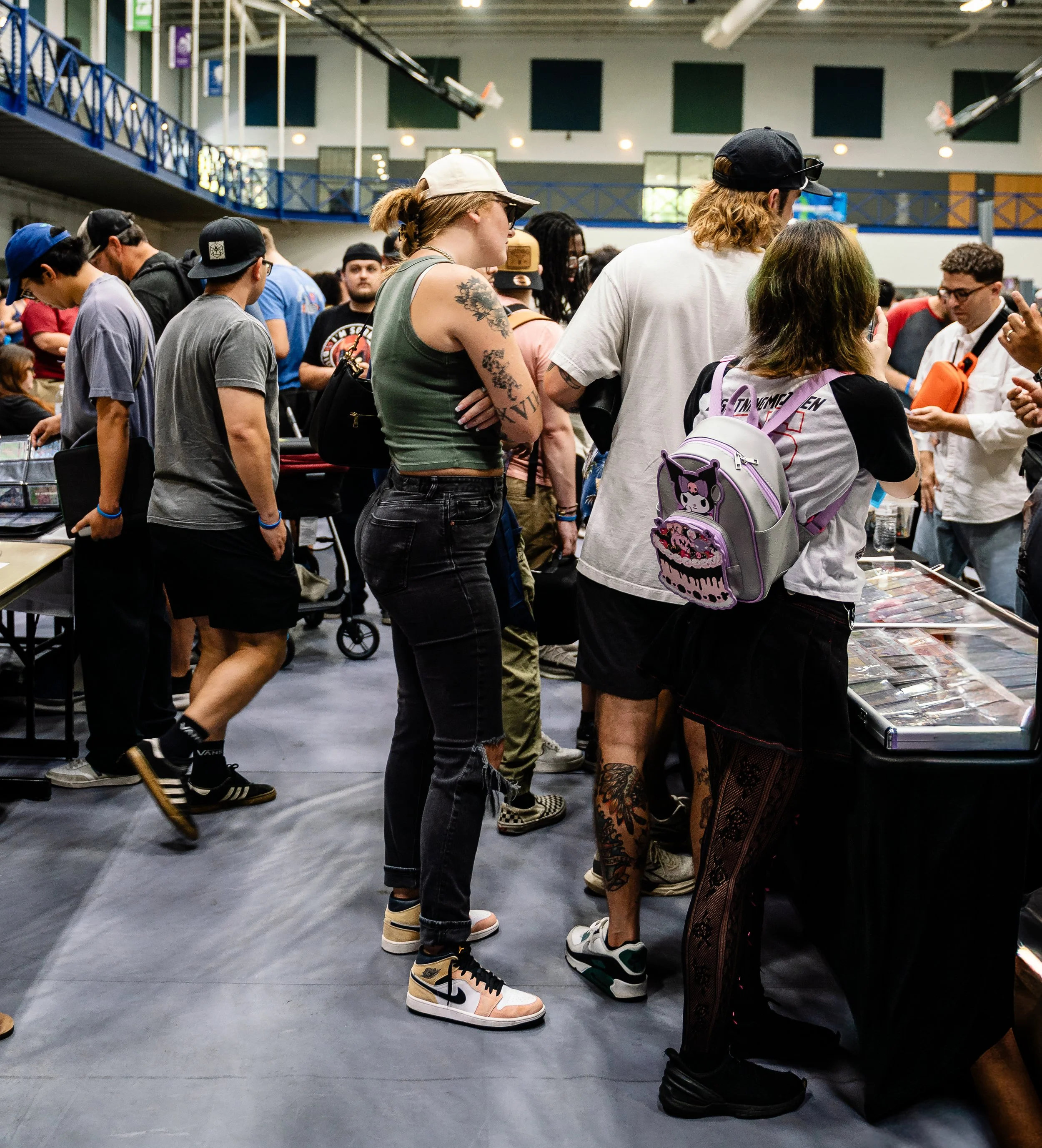 A group of people in line at an indoor event, browsing merchandise table, some wearing casual clothes and backpacks, with various hairstyles and tattoos, in a large warehouse-like space.