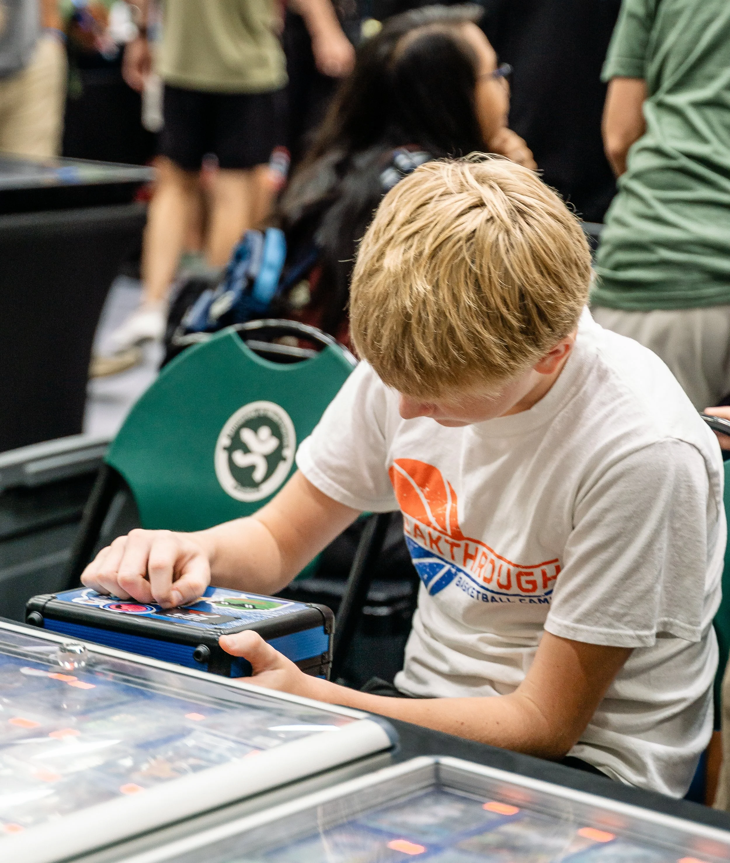 A young boy with blonde hair and a baseball t-shirt playing a tabletop arcade game at an event, with colorful game cards and blurred people in the background.