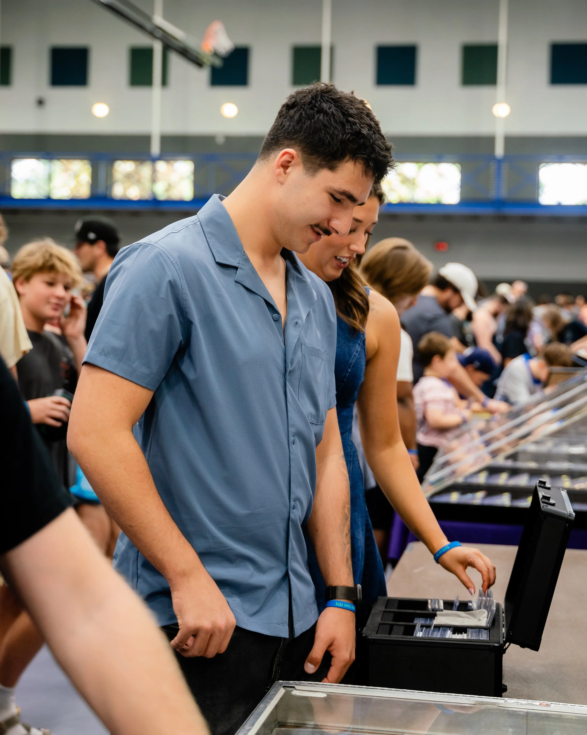 A young man with short curly hair wearing a blue shirt, looking down at a display case at an indoor event, with other people in the background.