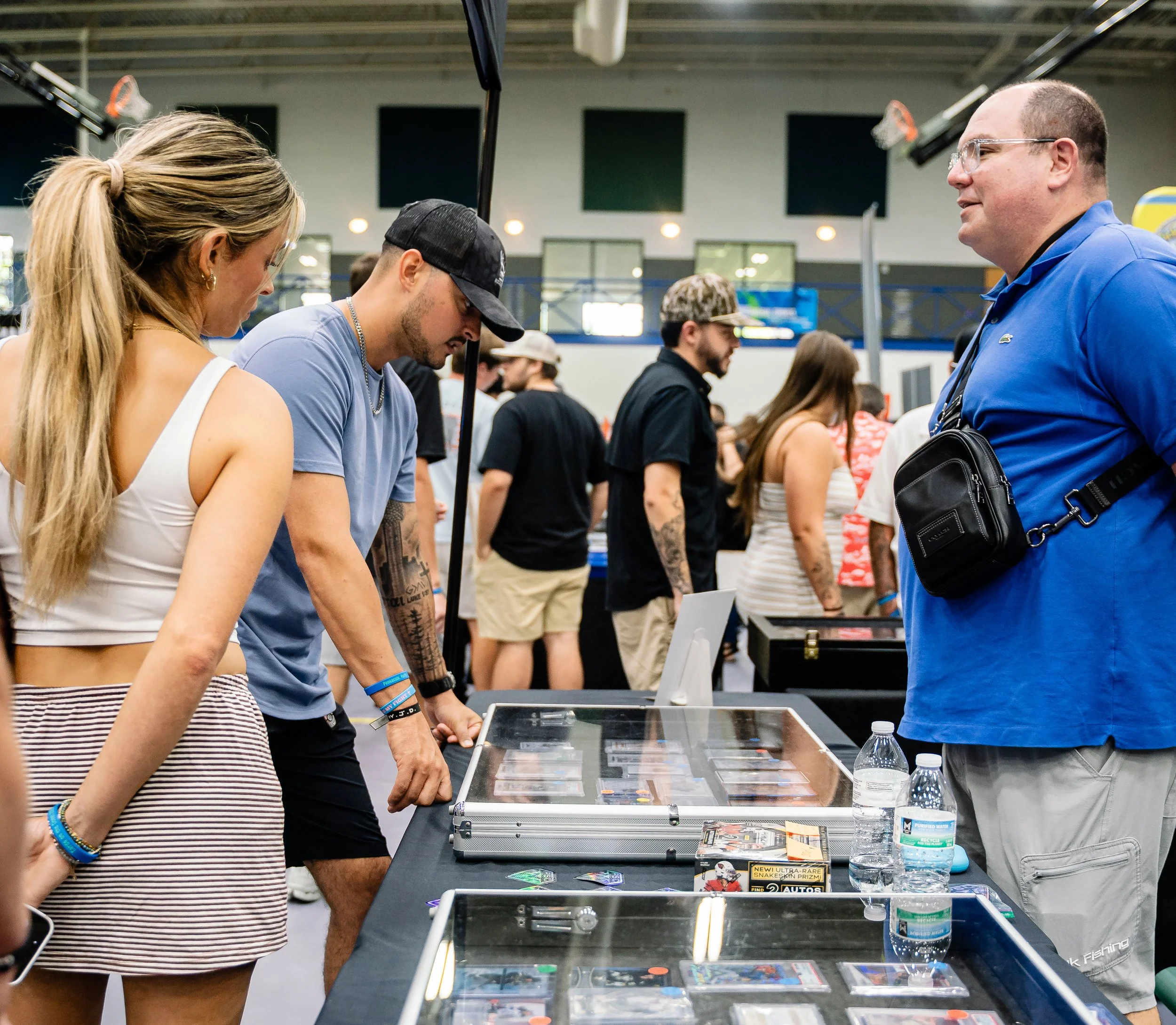 People at a gaming or trading event, closely examining items on a display table, with others in the background in a large indoor venue.
