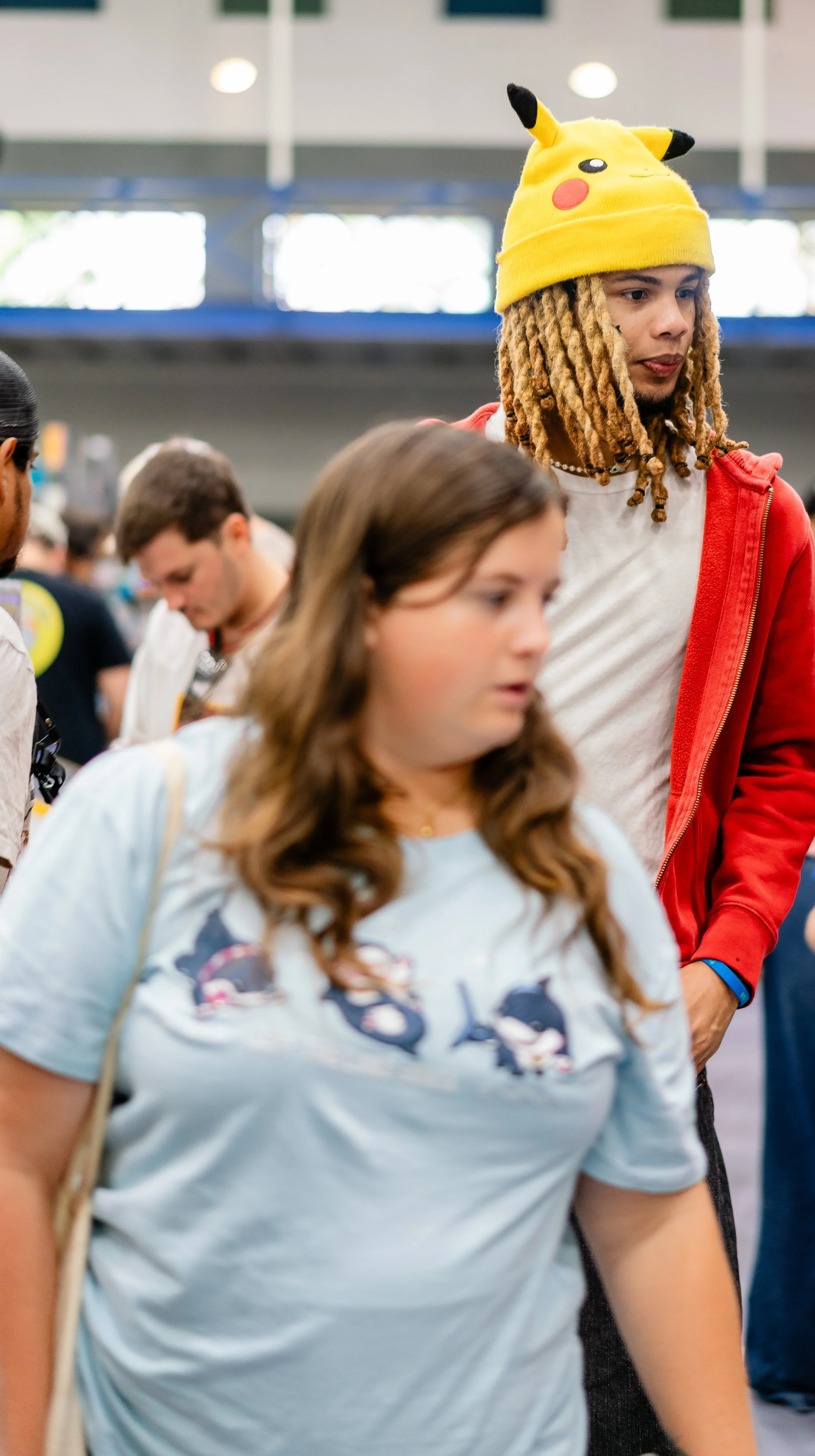 A young man with dreadlocks wearing a Pikachu hat and a red jacket, standing in a crowded indoor space, with a young woman in the foreground and other people in the background.