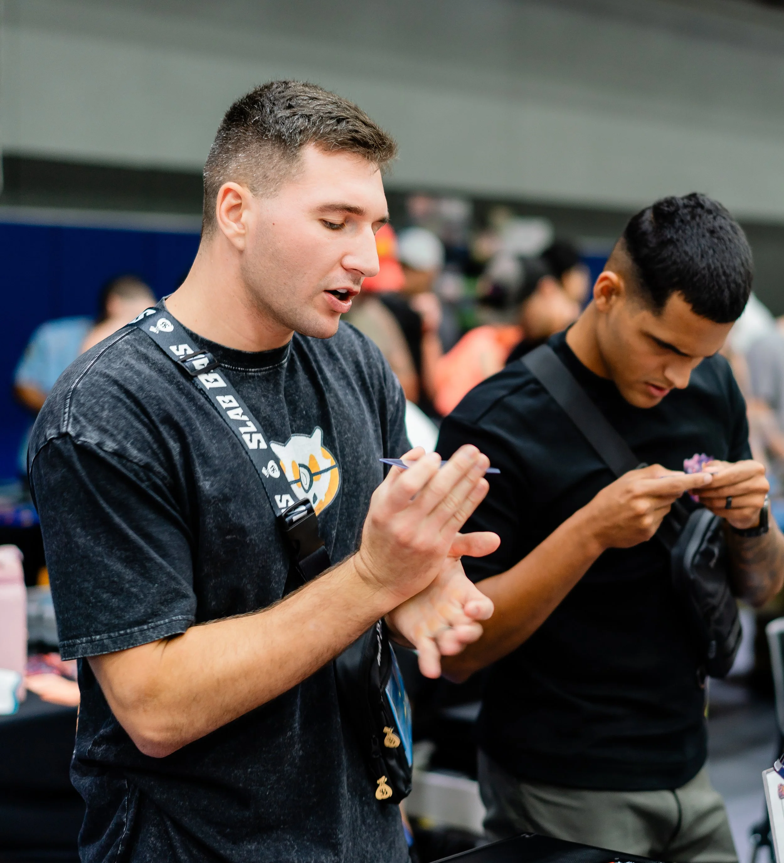 Two people are seen from the shoulders up, engaged in a conversation or activity, with the man on the left holding a card or paper, in an indoor setting with blurred background of other people.