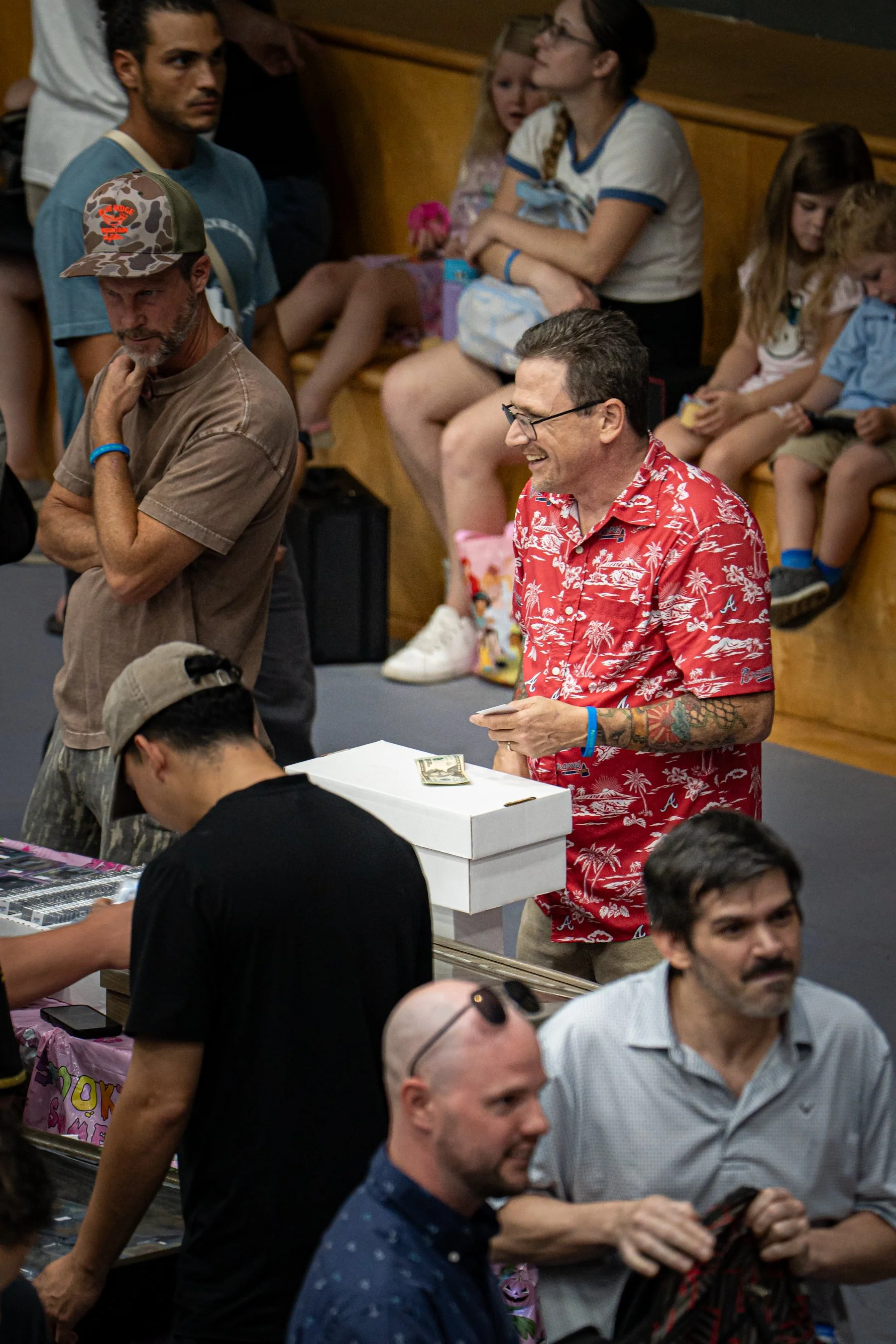 A group of people at a convention or event, some standing and some seated, with tables and boxes around them.