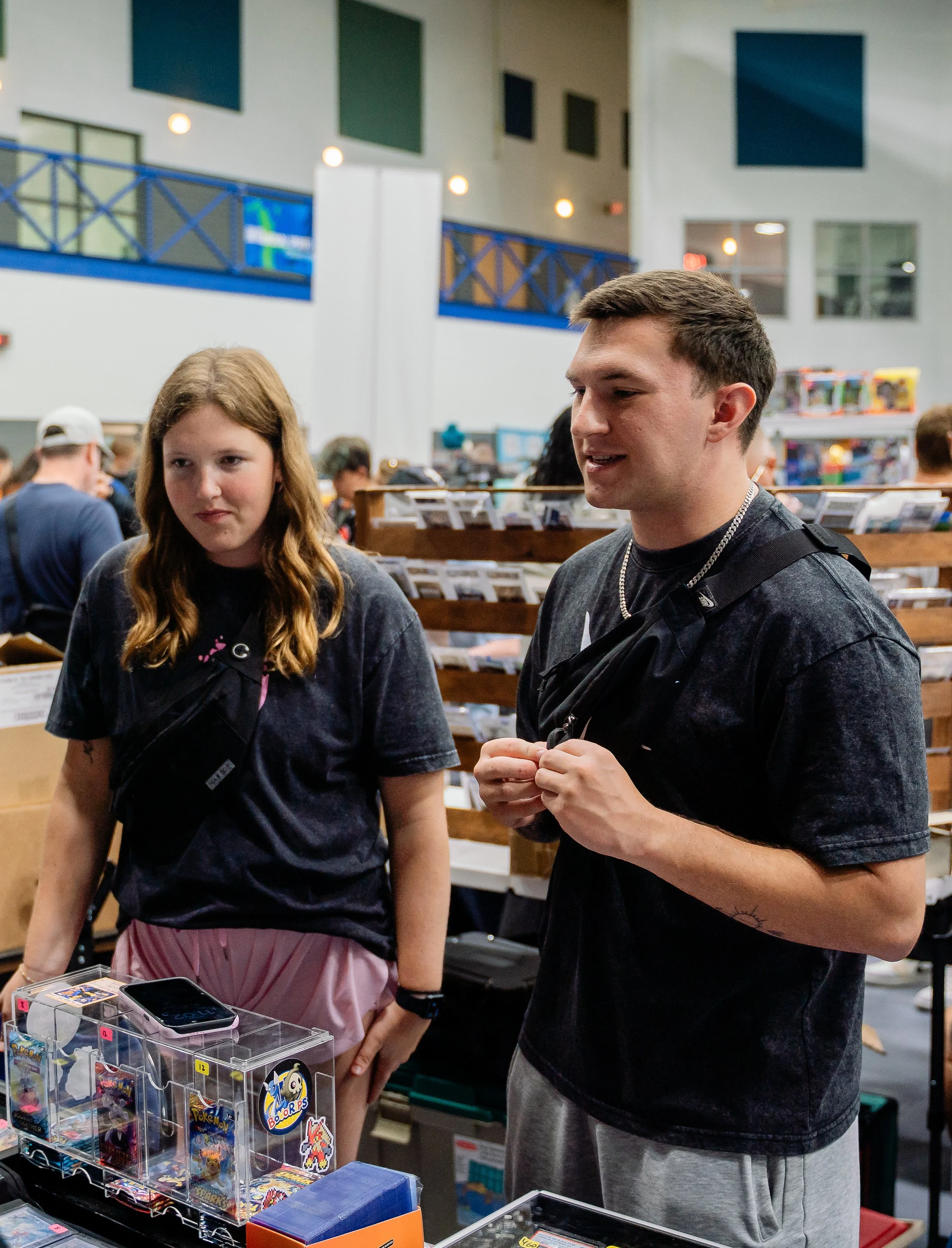 A young man and a young woman browsing Pokémon trading cards at a store, with shelves of collectibles and gaming items behind them.