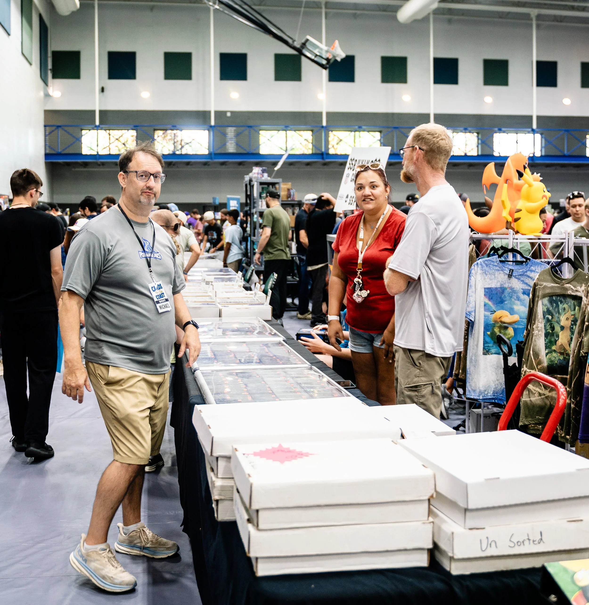 Man in gray shirt and shorts standing next to a table with unorganized items at a crowded indoor event, and a woman in red talking to a man with glasses. There are balloons and other attendees in the background.