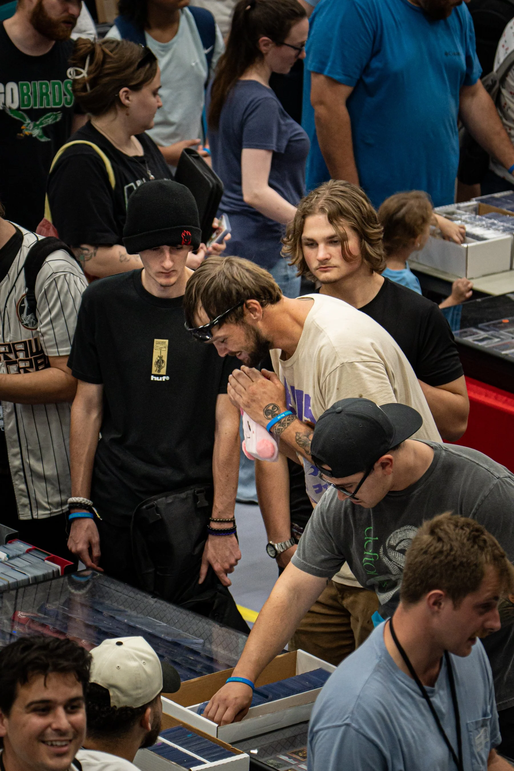 People shopping at a comic or trading card convention, browsing tables with trading cards, action figures, and collectibles.