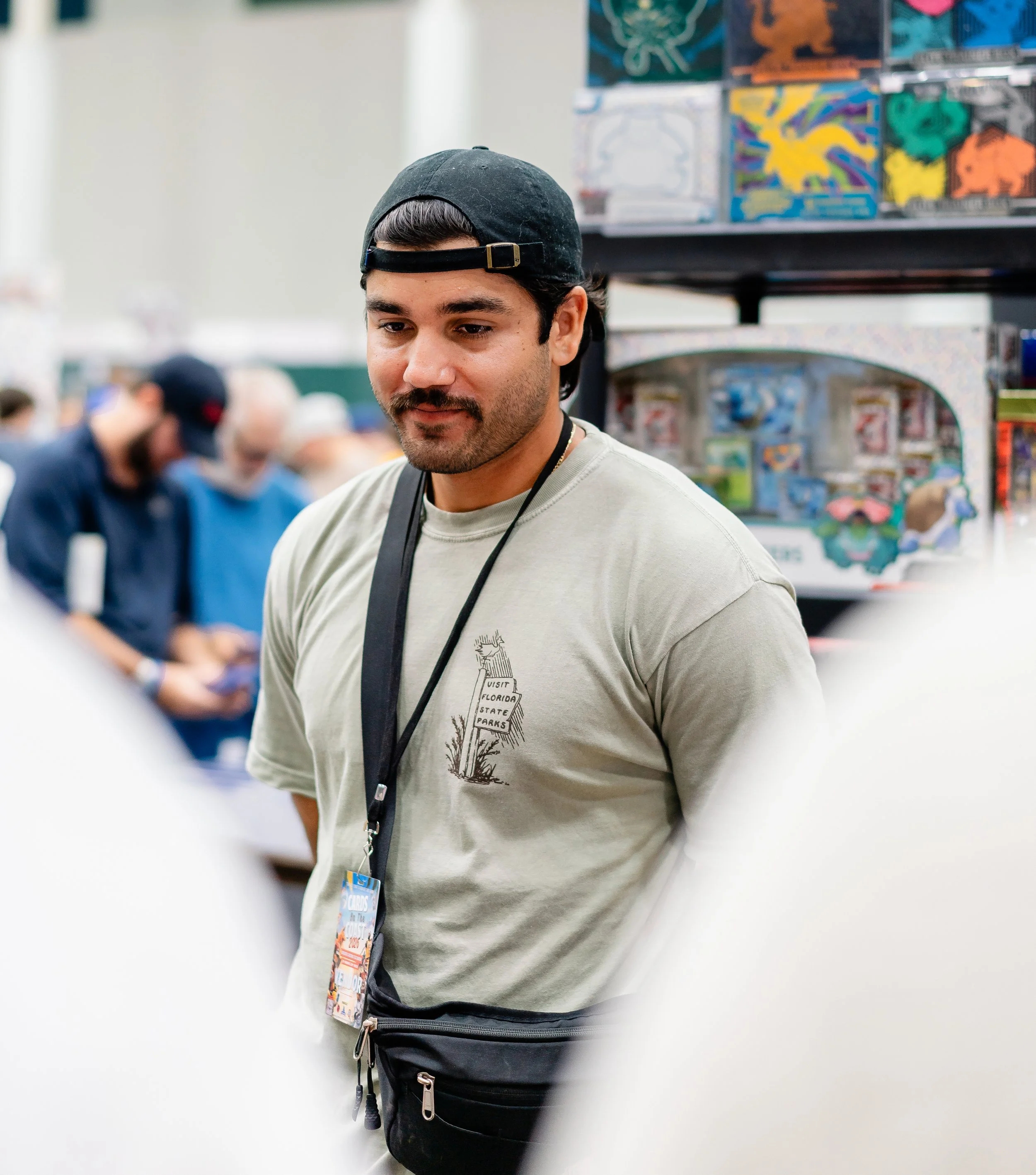 A young man with dark hair and facial hair wearing a backward baseball cap, a light gray T-shirt with a graphic, and a black waist bag, standing in a store aisle with toys and games in the background.