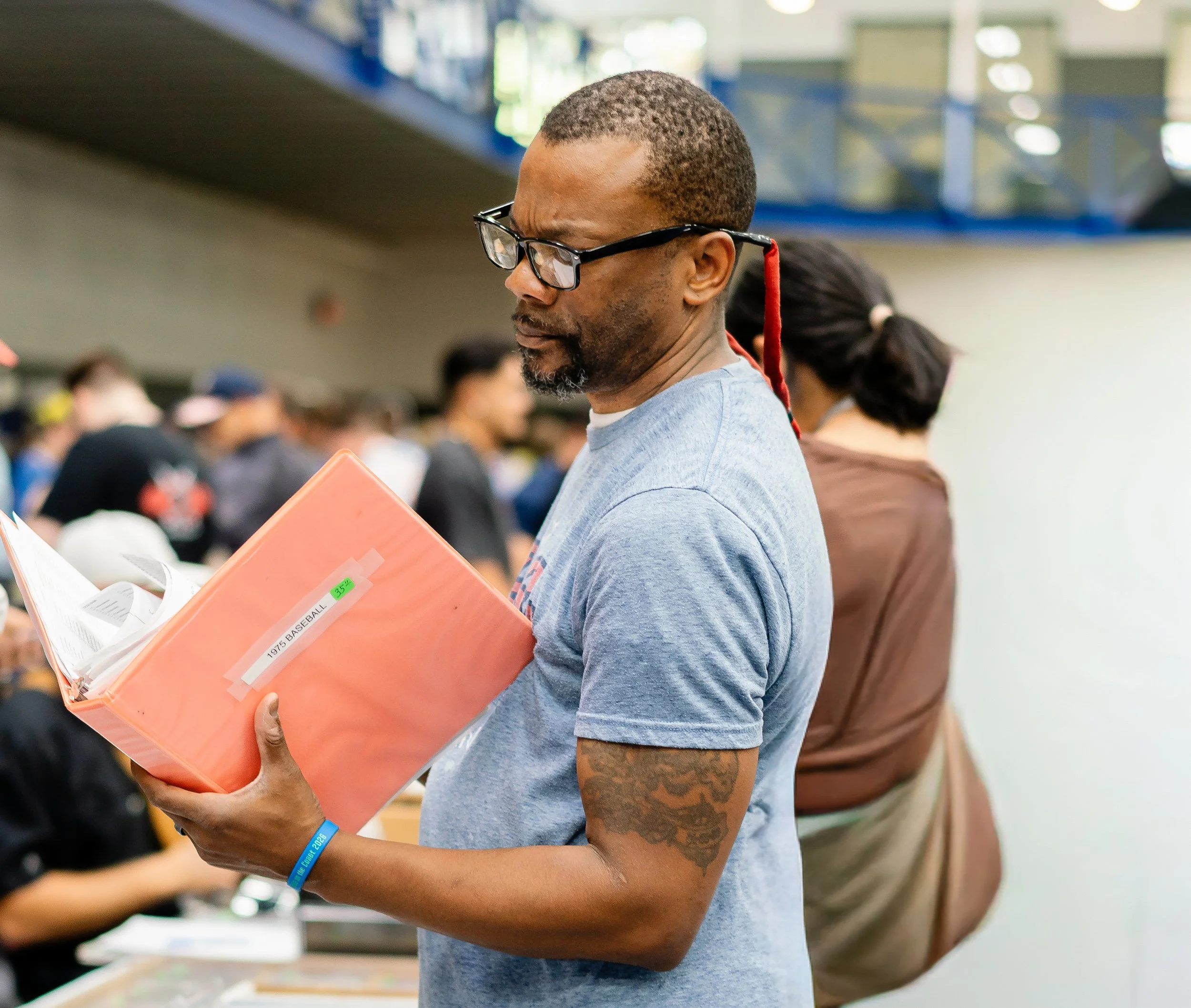 A man with glasses reading a book, holding a pink folder, at an indoor event with other people in the background.