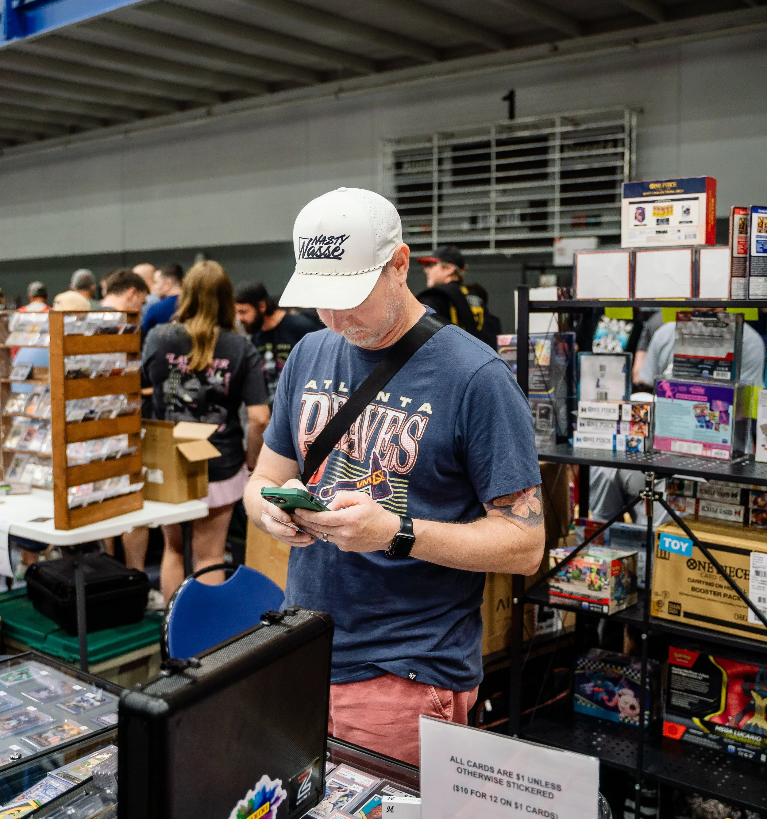 Person wearing a white baseball cap with the words "Nasty Nate" printed on it, shopping at a game store, with various trading cards and merchandise around.